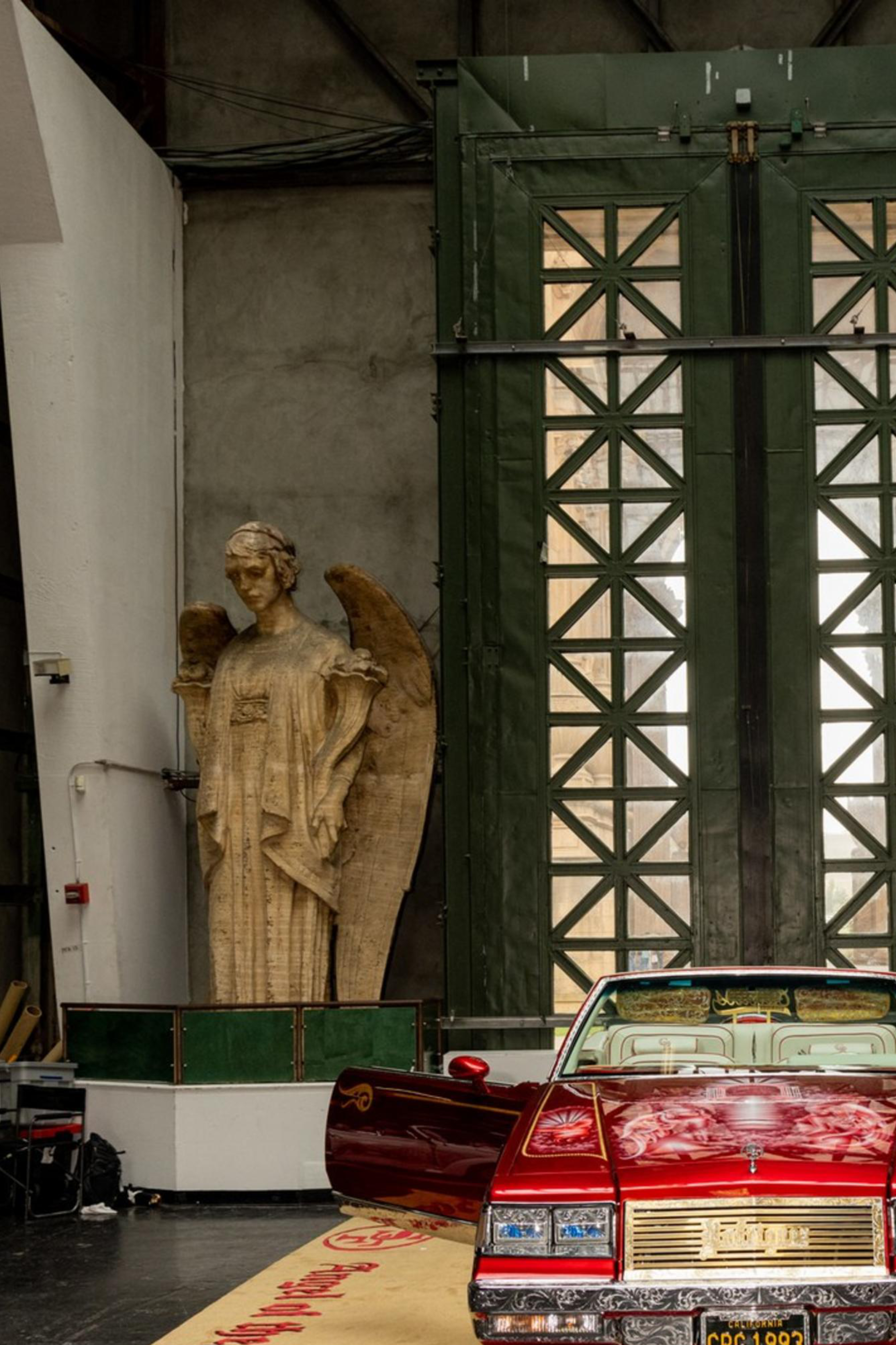 The image shows a vintage car exhibition with a red convertible in the center on a carpet, flanked by white and mint-green cars. Two large angel statues are in the background.
