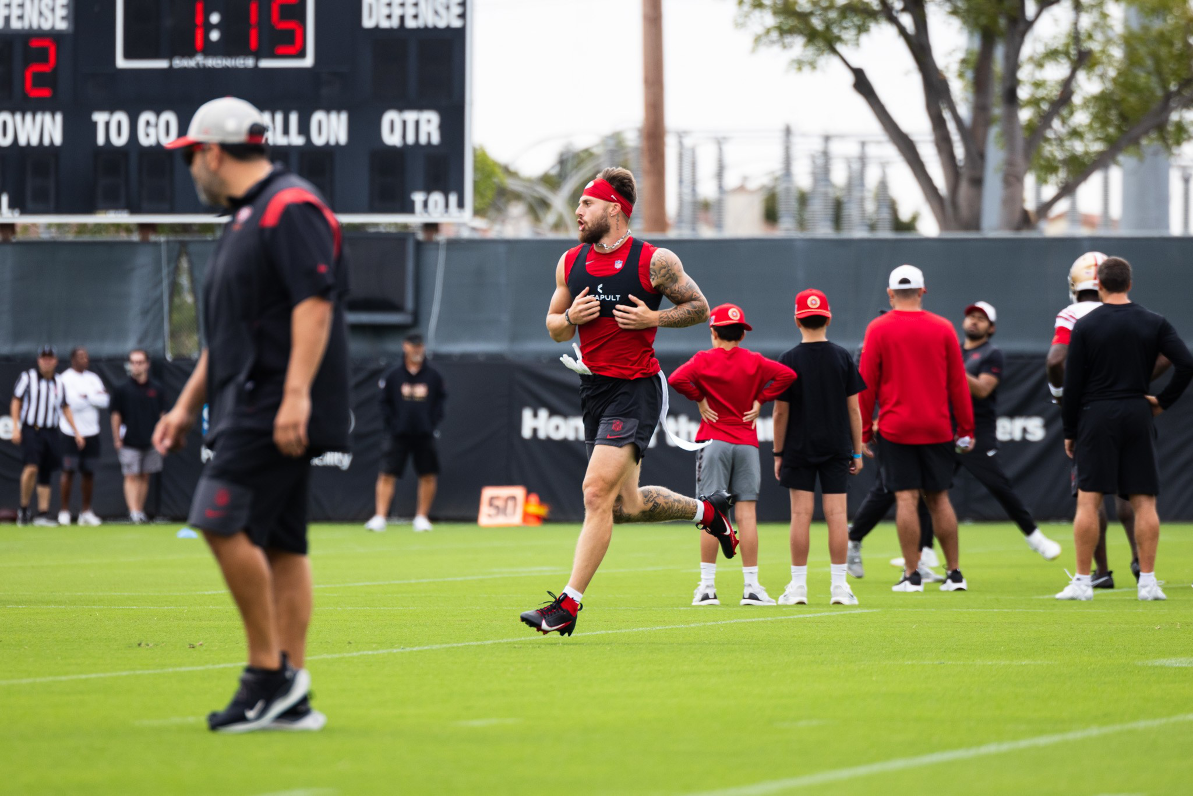 A football player in a red vest runs on a grass field, watched by coaches and young fans. A digital scoreboard and trees are visible in the background.