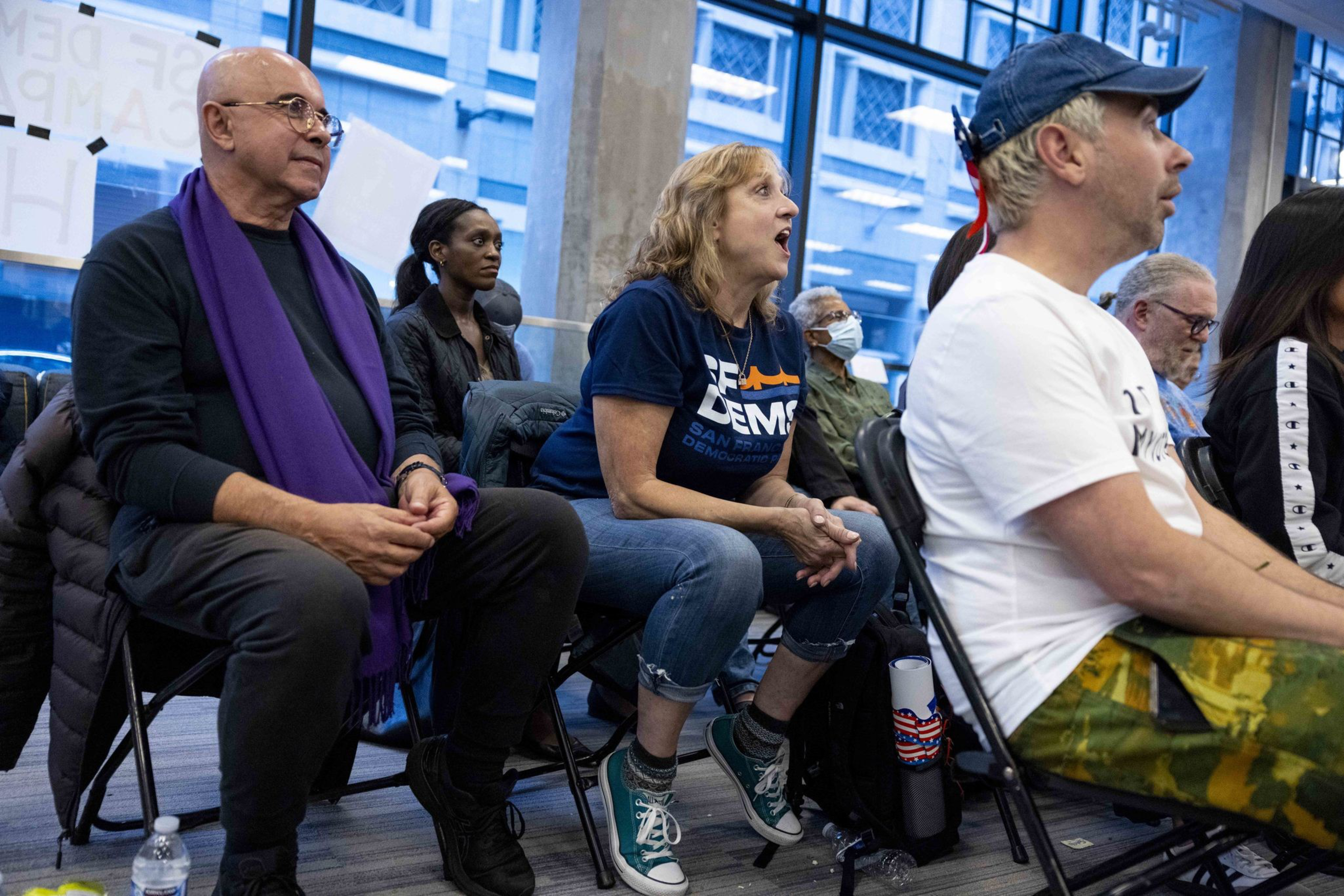 A group of people sits attentively in a room with large windows. One person wears a &quot;Dems&quot; t-shirt and another sports a purple scarf. Some wear masks, and all are focused.