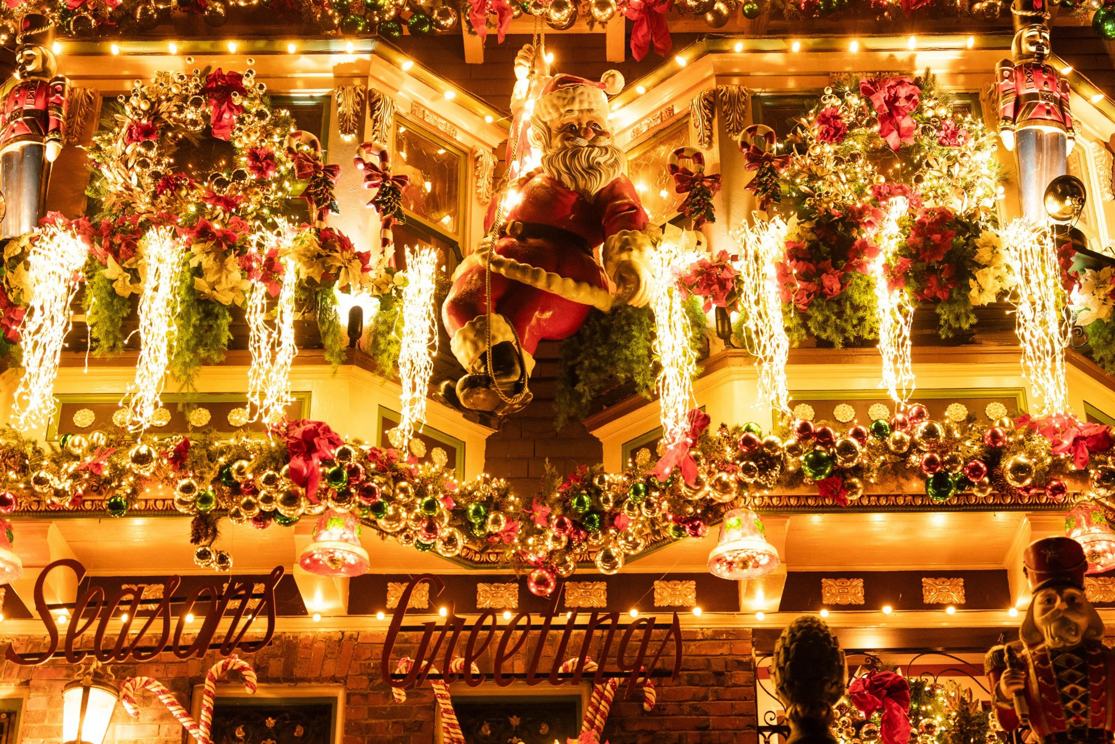 A festive building is adorned with a Santa figure, bright lights, wreaths, garlands, and nutcracker decorations, with &quot;Seasons Greetings&quot; displayed below.