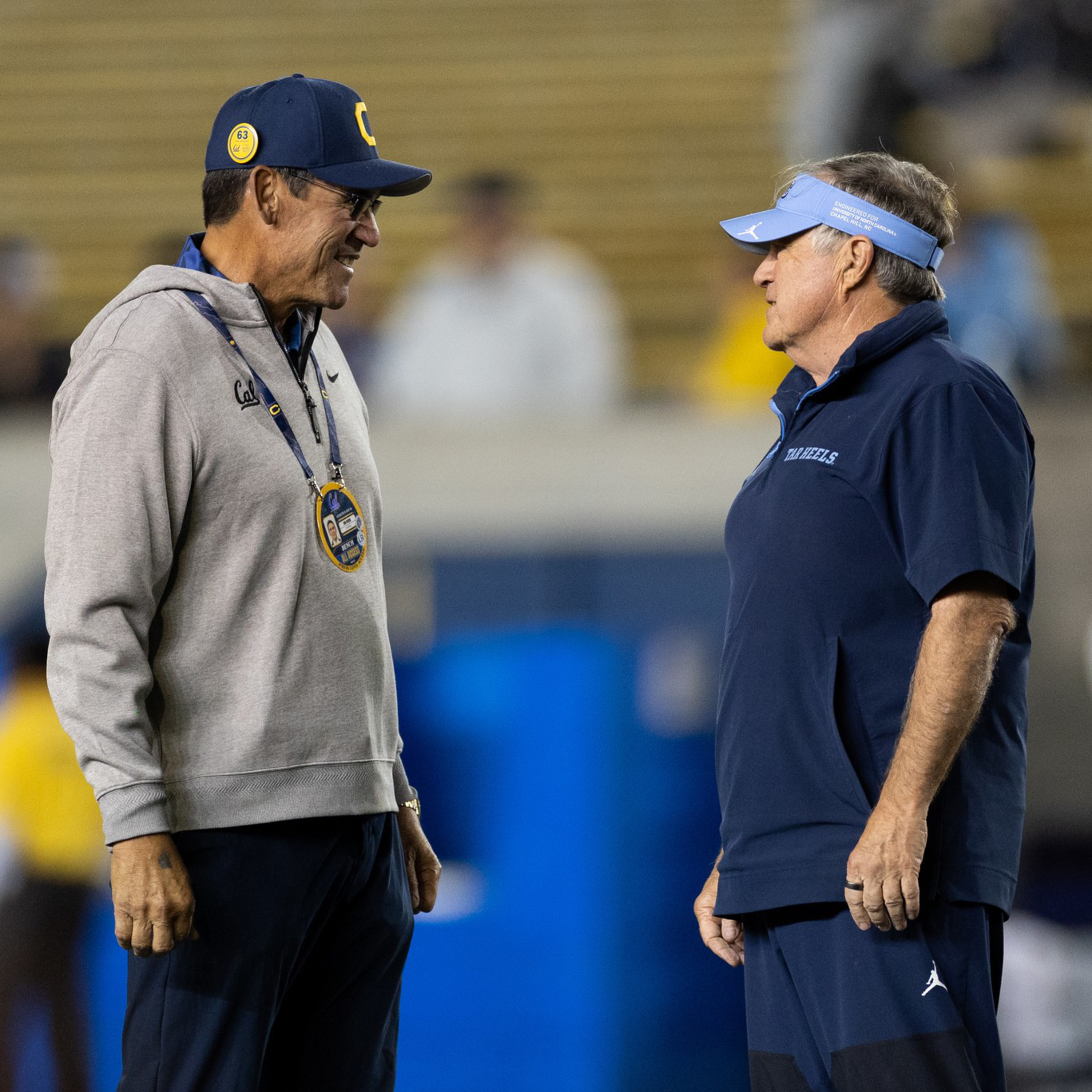 Two men, one in gray and blue with a stopwatch and cap, and the other in dark blue with a visor, stand facing each other in conversation on a field.