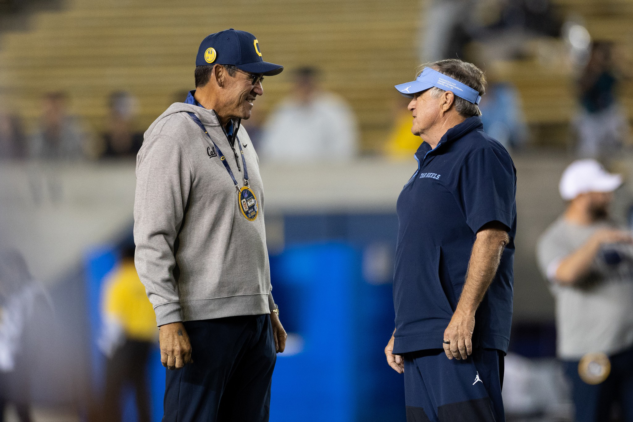 Two men, one in gray and blue with a stopwatch and cap, and the other in dark blue with a visor, stand facing each other in conversation on a field.