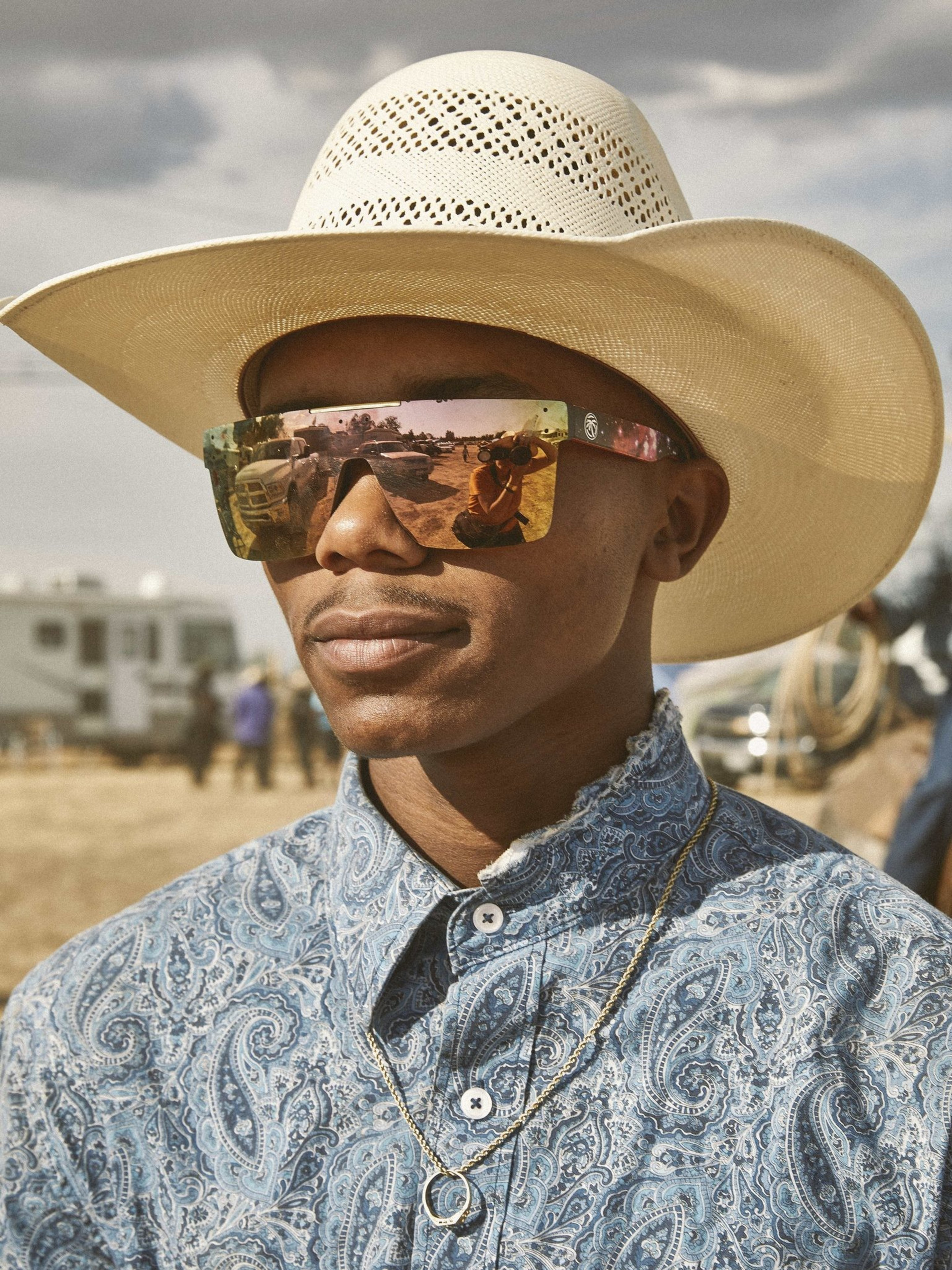 A person wearing a light-colored cowboy hat, reflective sunglasses, and a paisley-patterned shirt stands outdoors, with a trailer and a dusty field in the background.