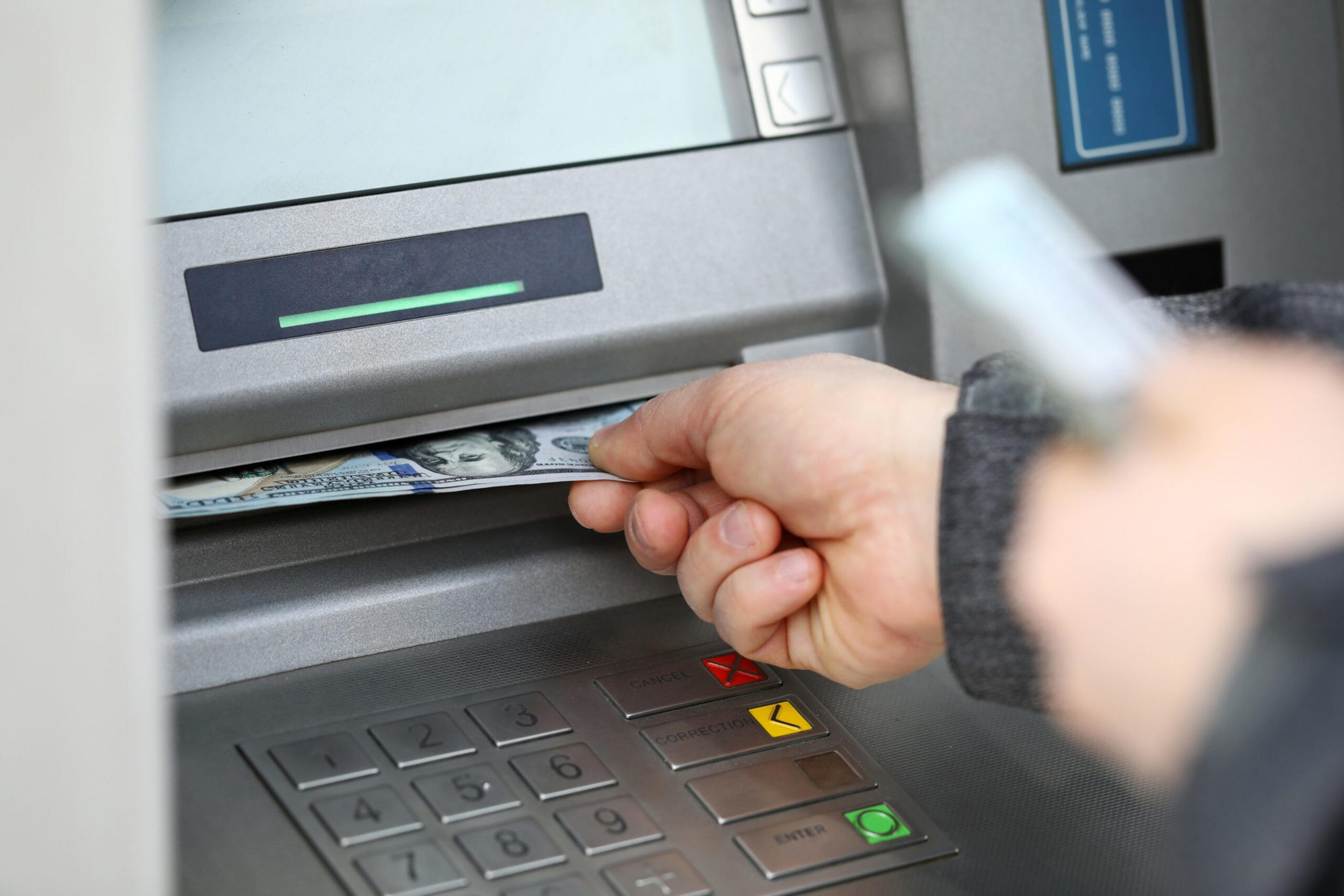 A hand is pulling out US $100 bills from an ATM, with the keypad and card slot visible nearby.
