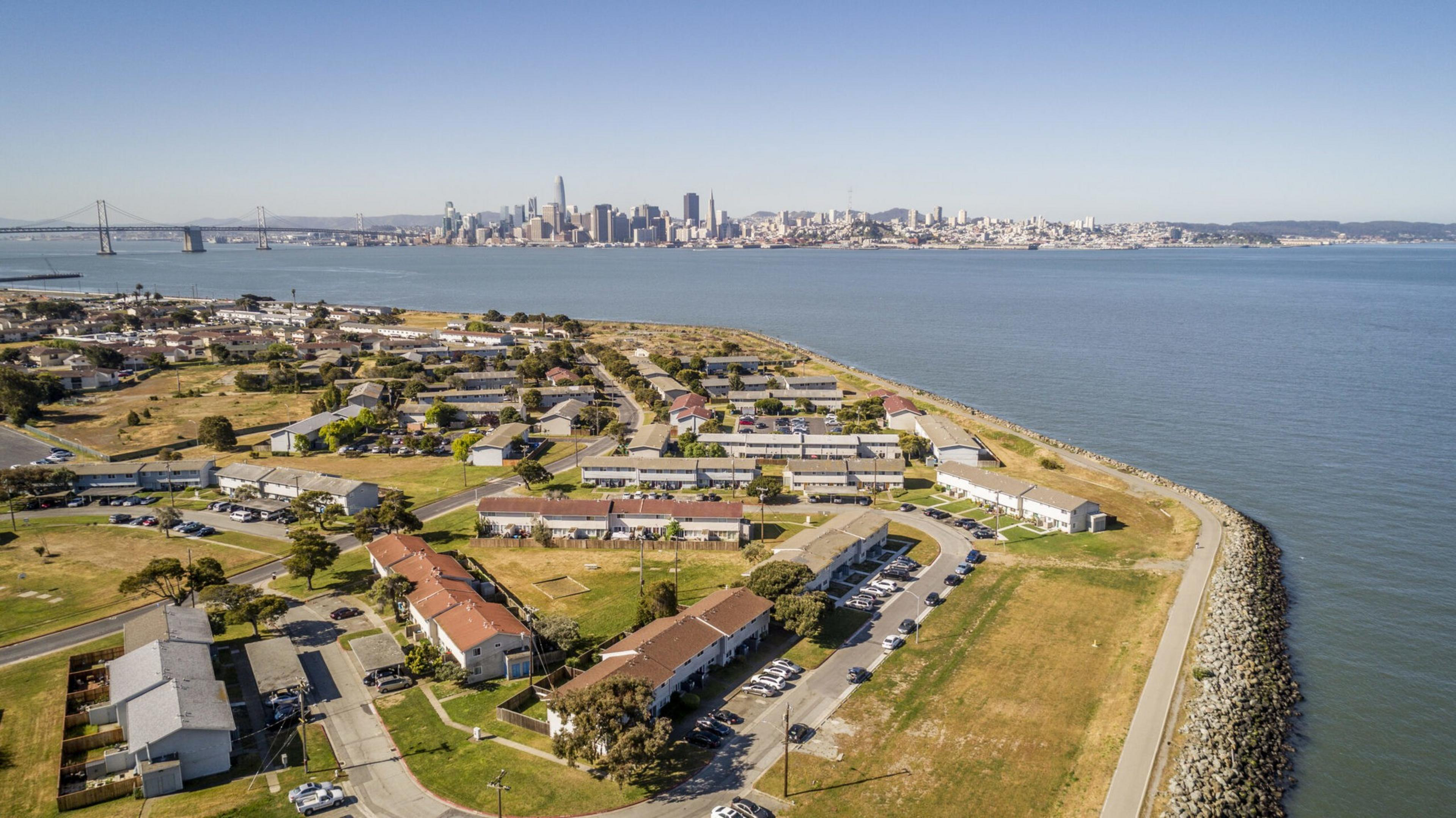 A coastal residential area with houses and parking lots borders a calm bay, with a distant city skyline and a large suspension bridge across the water.
