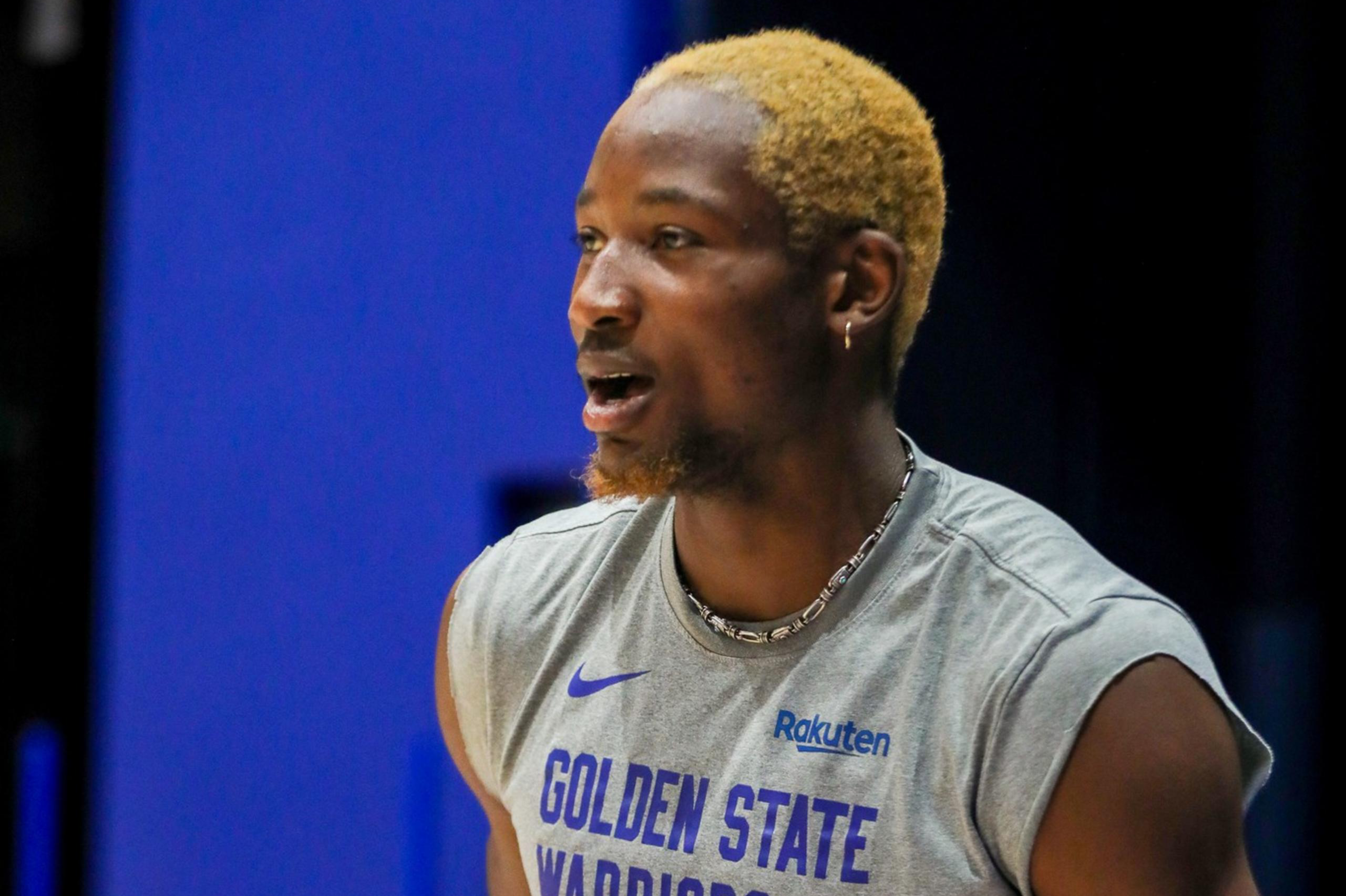 A basketball player with bleached hair holds a ball, wearing a gray "Golden State Warriors Basketball" sleeveless shirt, with a blue background behind him.