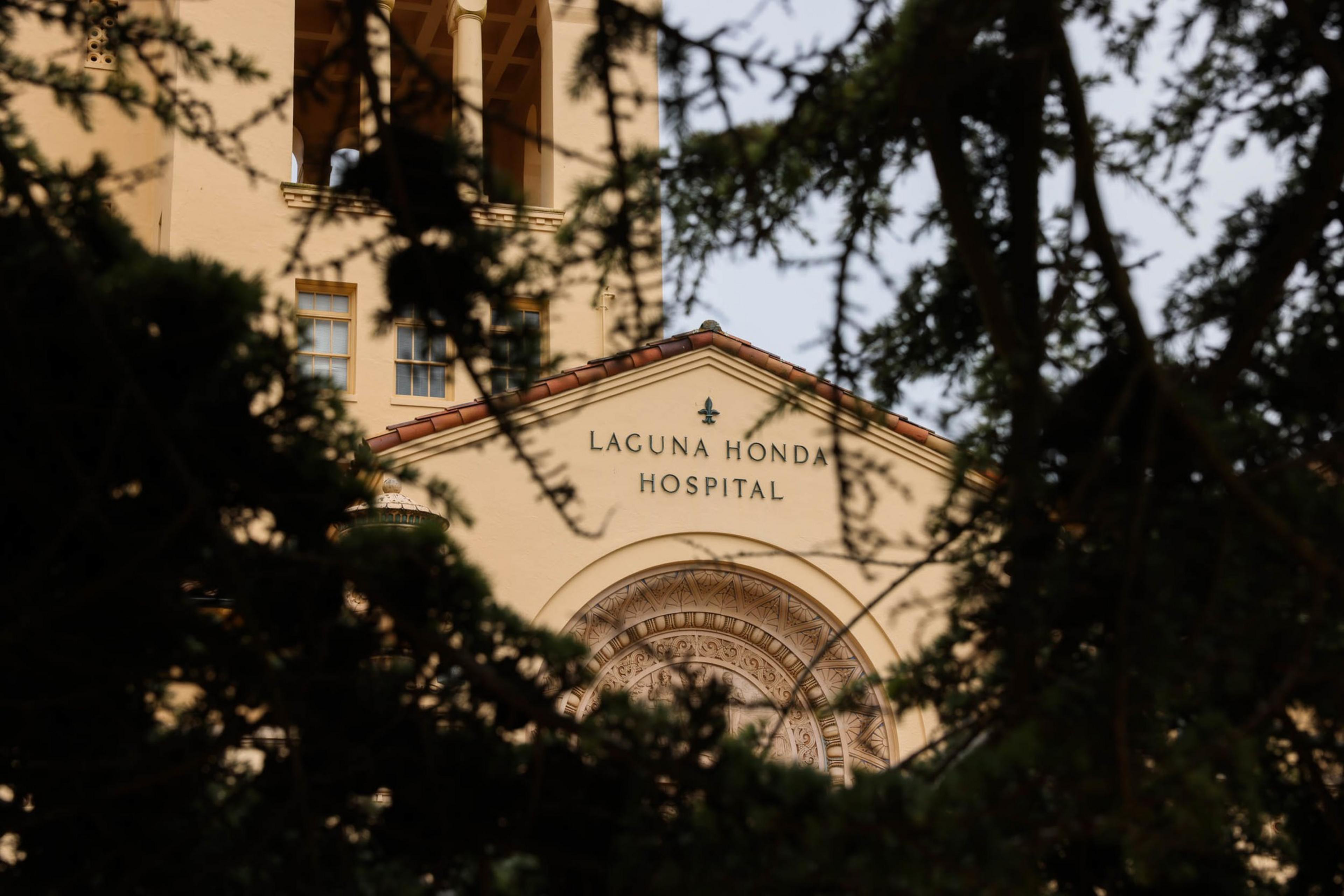 A beige building with "Laguna Honda Hospital" written on it is partially visible through dark green tree branches.
