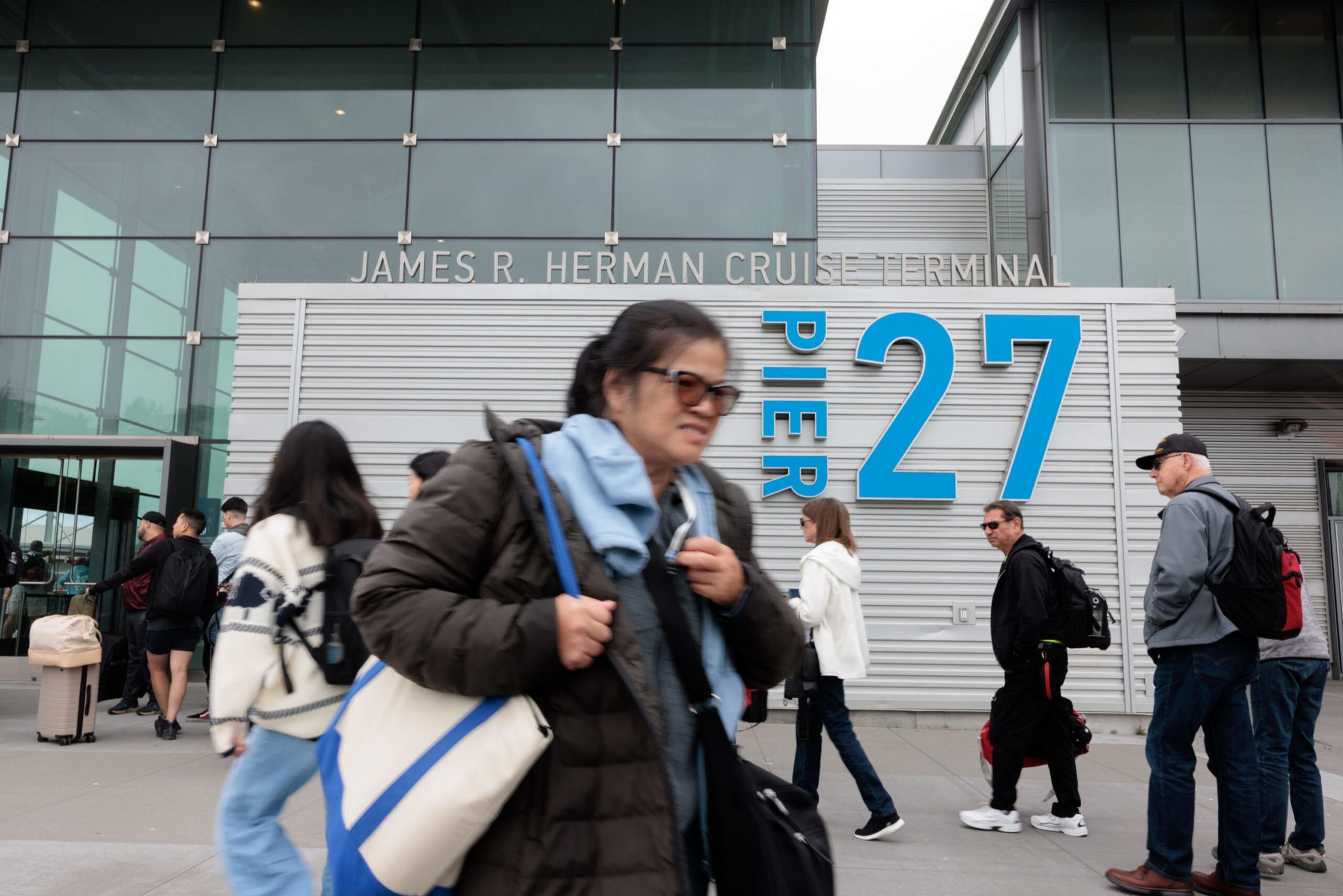 People walk past the James R. Herman Cruise Terminal at Pier 27, some carrying bags and dressed in jackets and casual wear.
