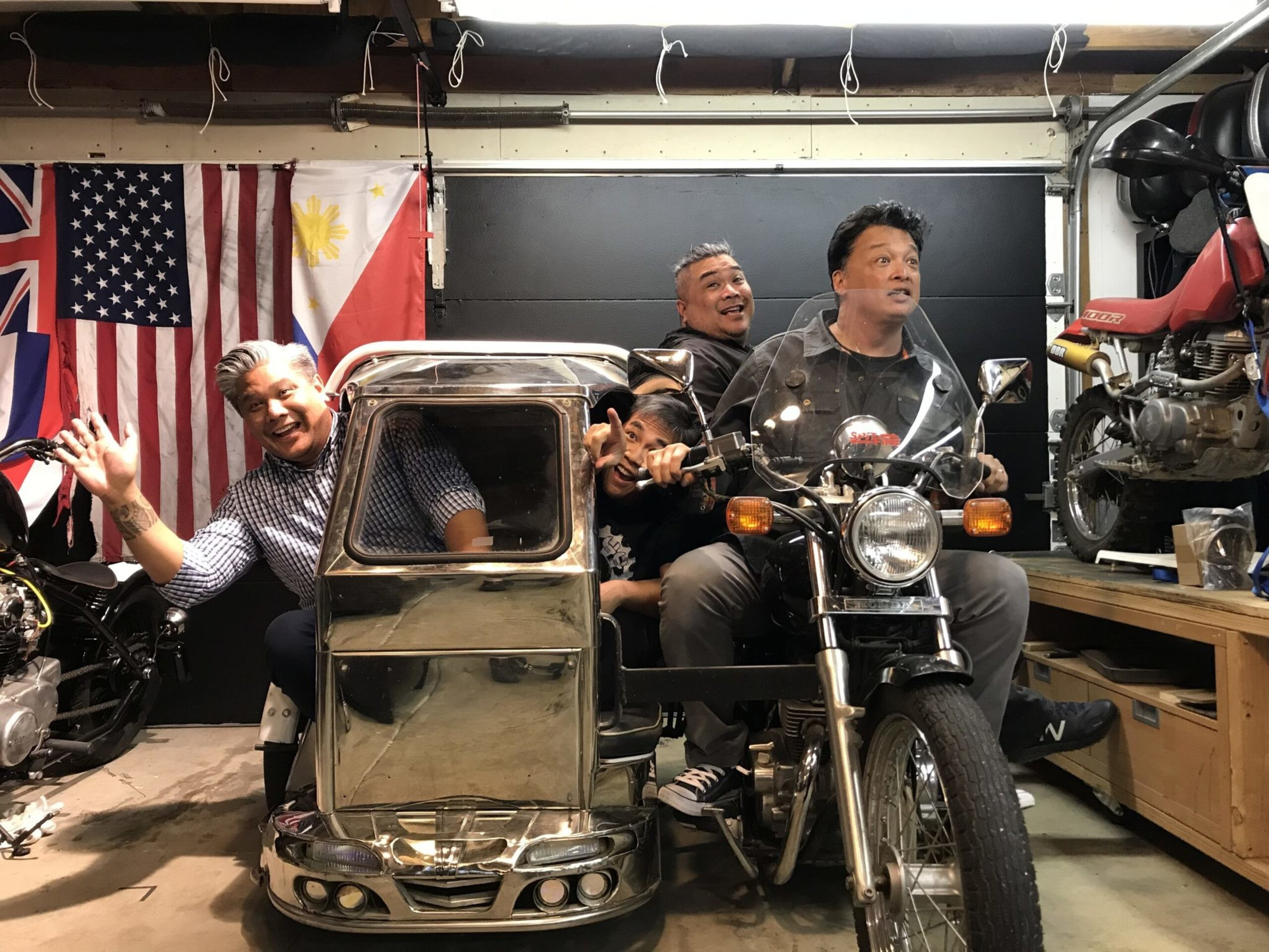 Four men smile and pose on a motorcycle with a shiny silver sidecar in a garage decorated with American, Hawaiian, and Filipino flags.