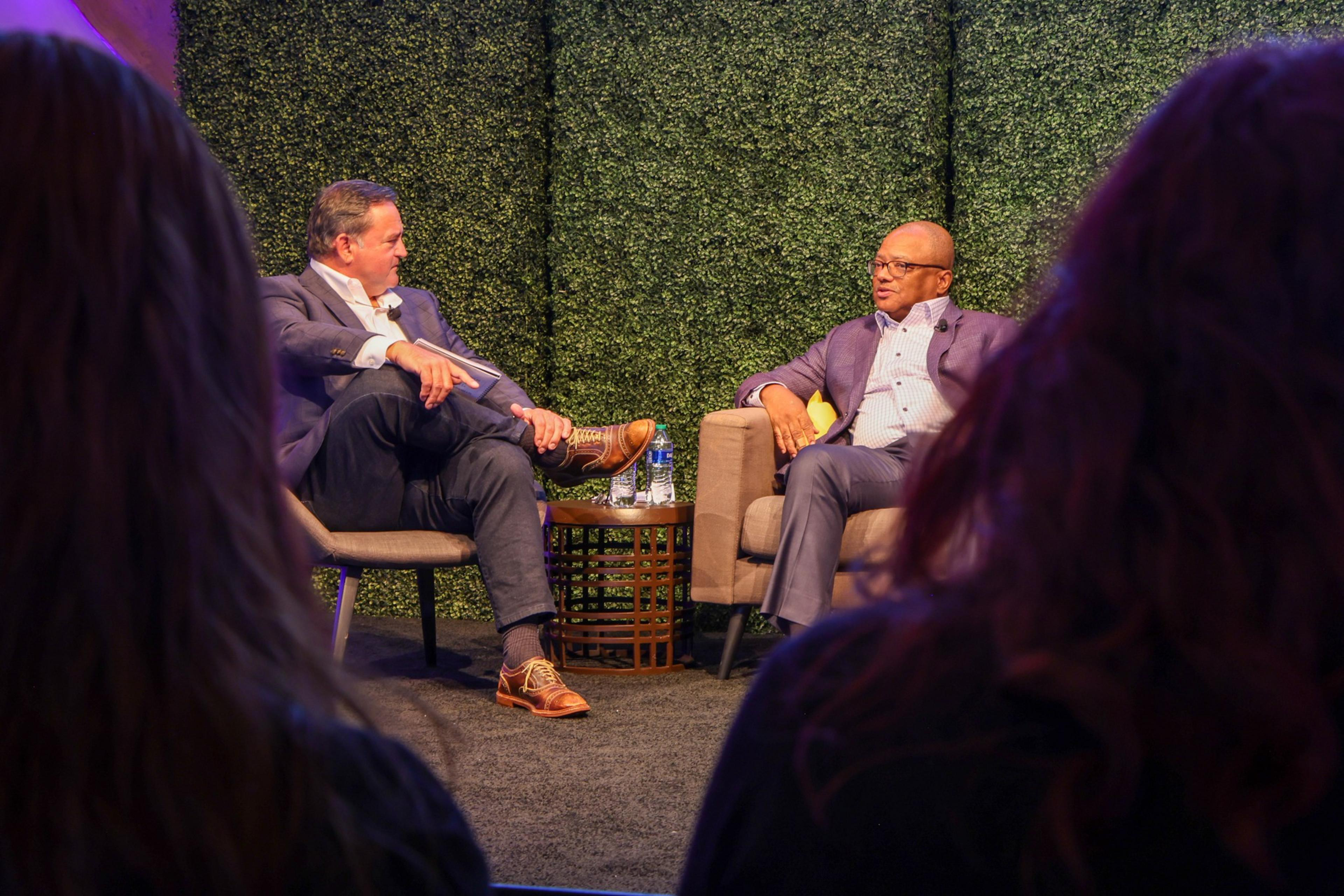 Two men are conversing on stage, seated in chairs, with an audience in the foreground and a green backdrop behind them.