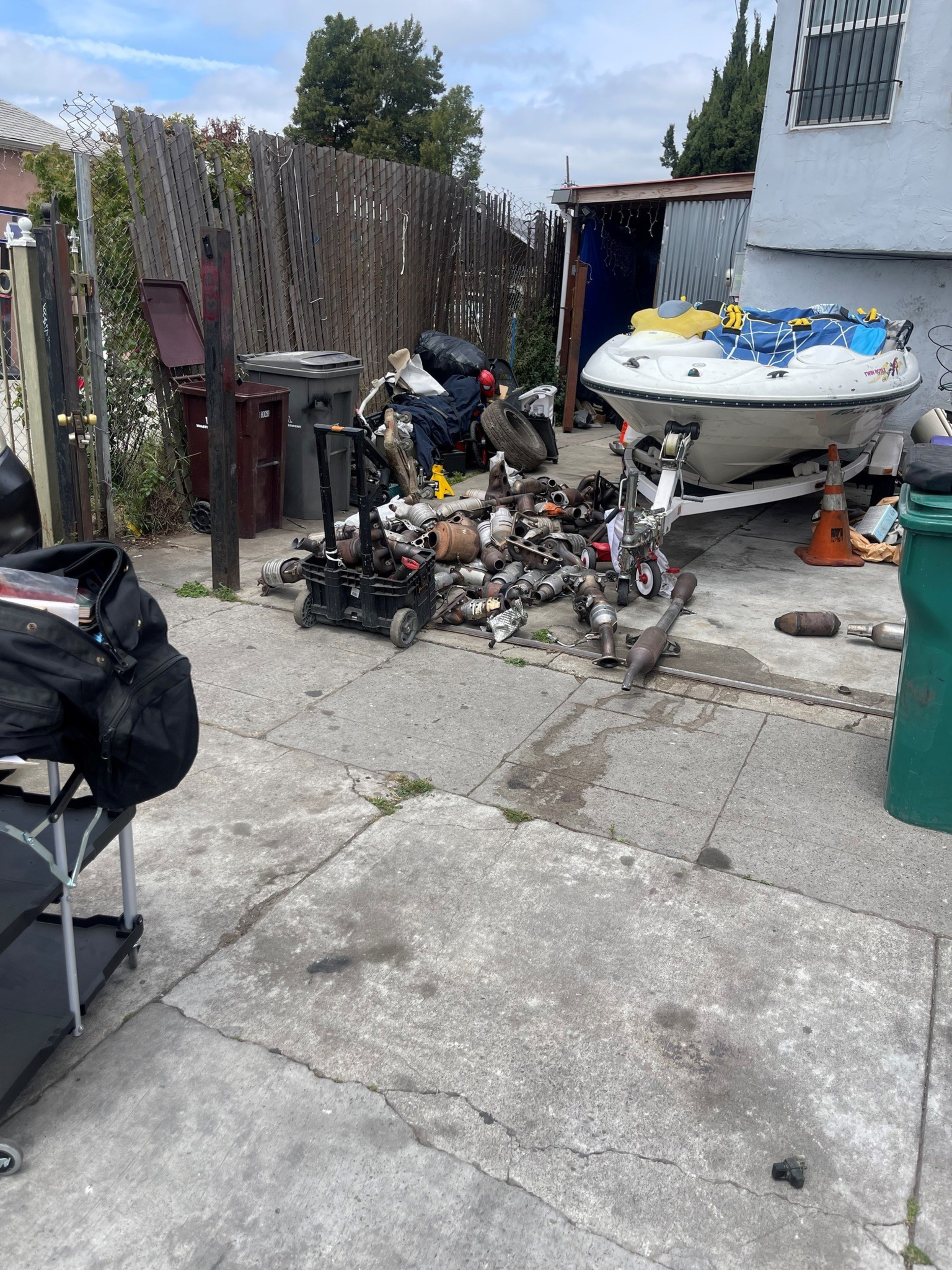 A small boat on a trailer sits next to a pile of metal parts and tools on a concrete driveway beside a wooden fence and trash bins.