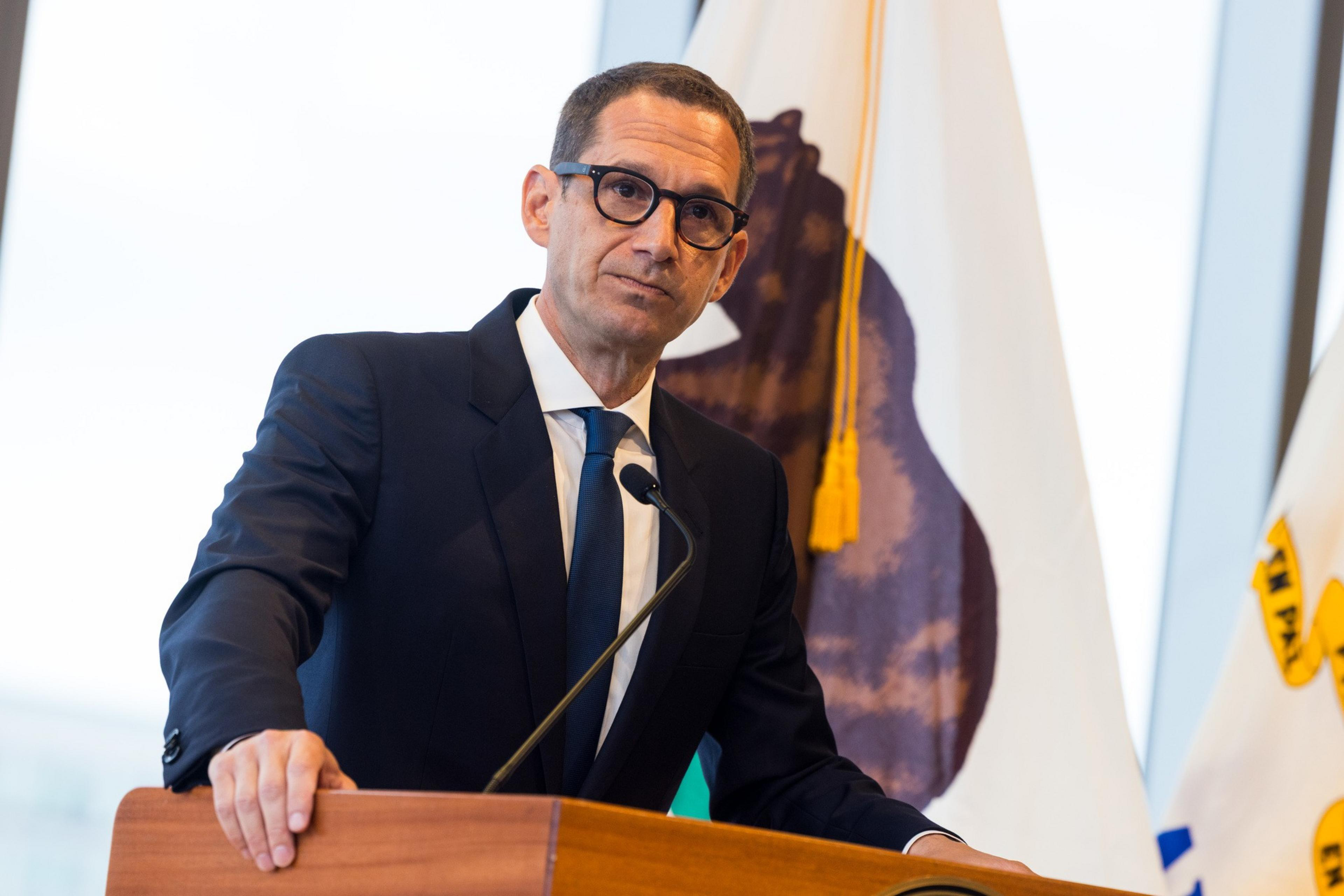 A man in a suit and glasses speaks at a podium with a microphone. Behind him are flags, one featuring a bear.