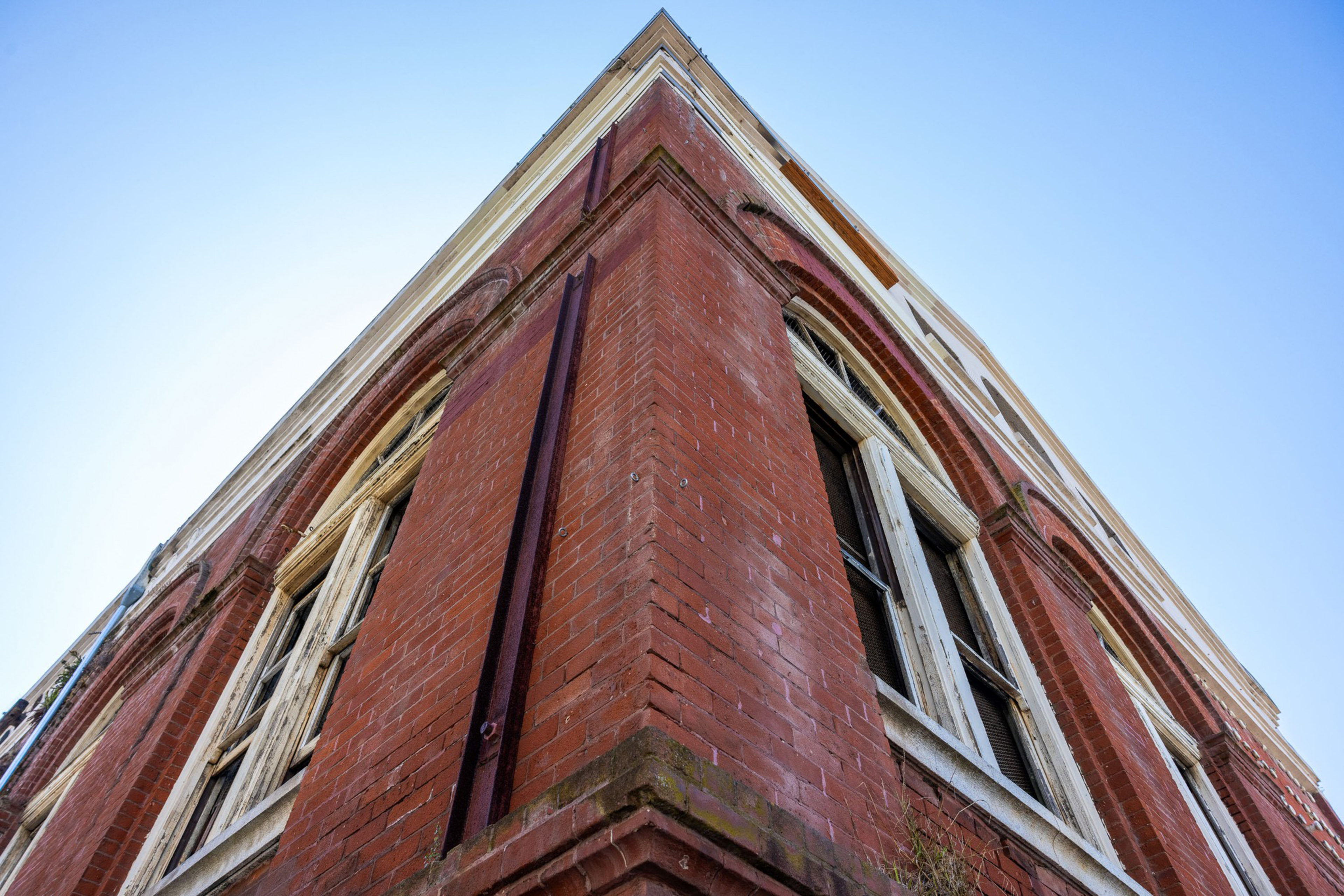 The image depicts the corner of a tall, red brick building with large, arched windows under a clear blue sky. The perspective is angled upwards, emphasizing its height.