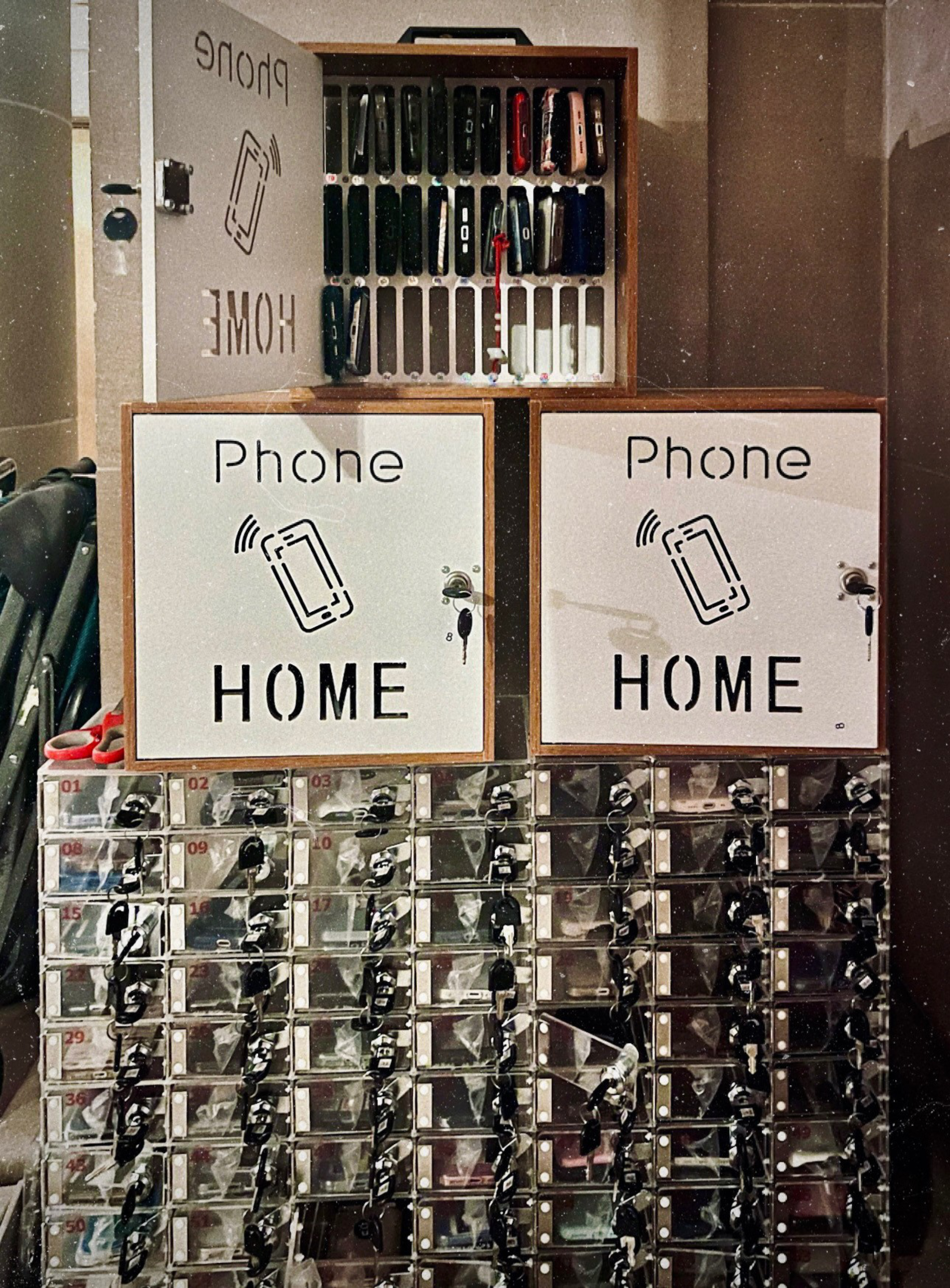 The image shows two boxes labeled &quot;Phone HOME&quot; with a charging station holding various phones on top. Below are small numbered lockers with keys attached.