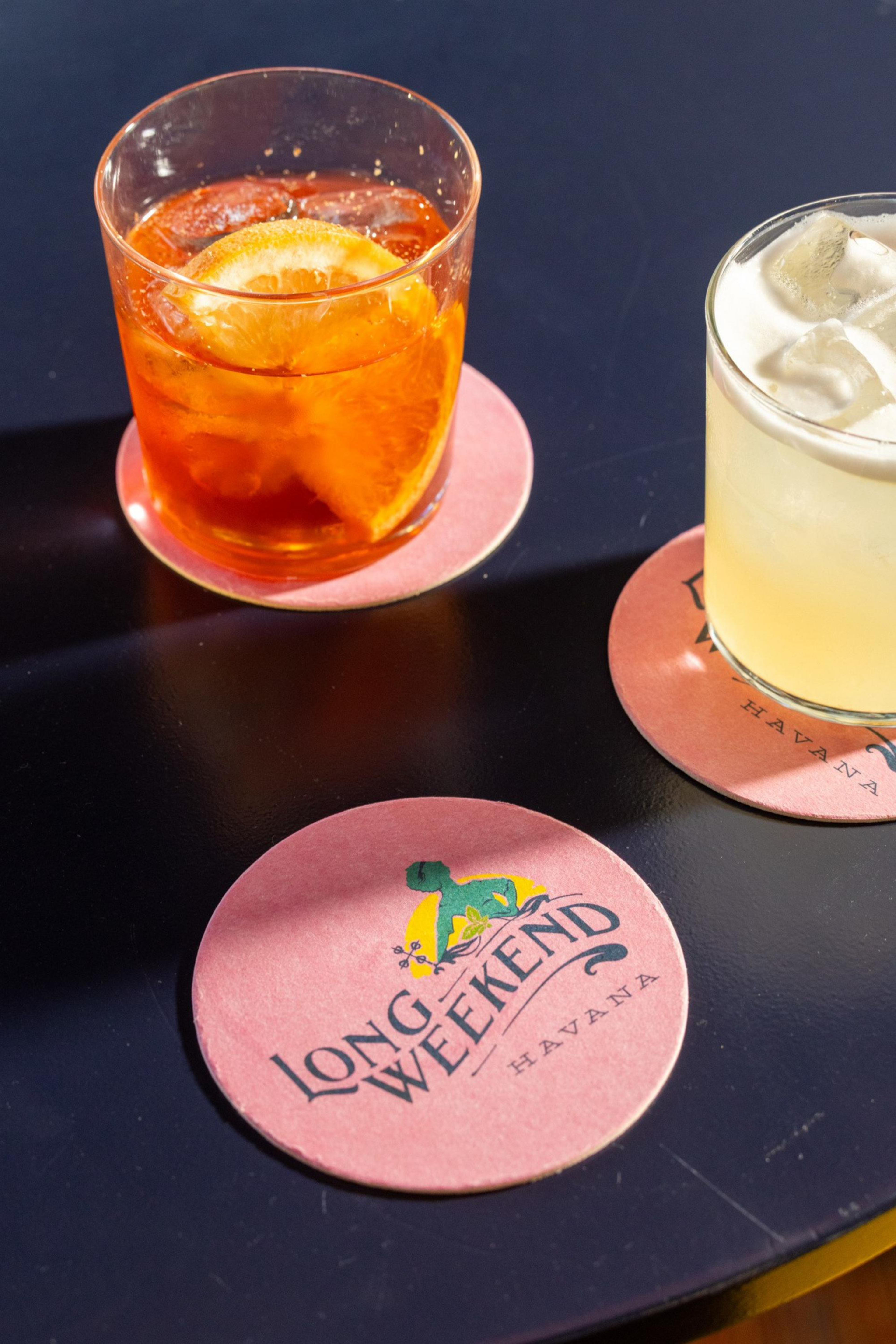 Two drinks on pink coasters labeled “Long Weekend Havana,” one orange with a lemon slice and ice, the other light yellow with ice, on a dark table.