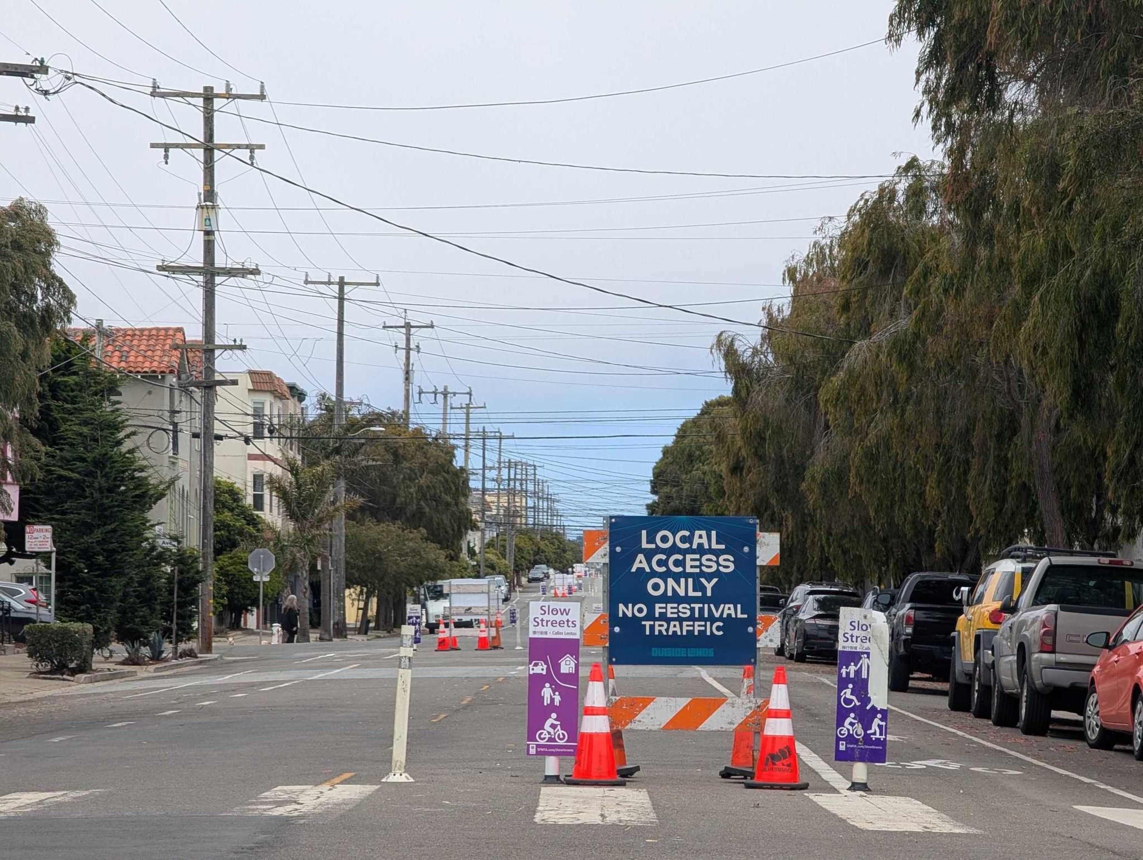 A street is partially blocked with traffic cones and signs stating "Local Access Only, No Festival Traffic" and "Slow Streets," with parked cars on the right.