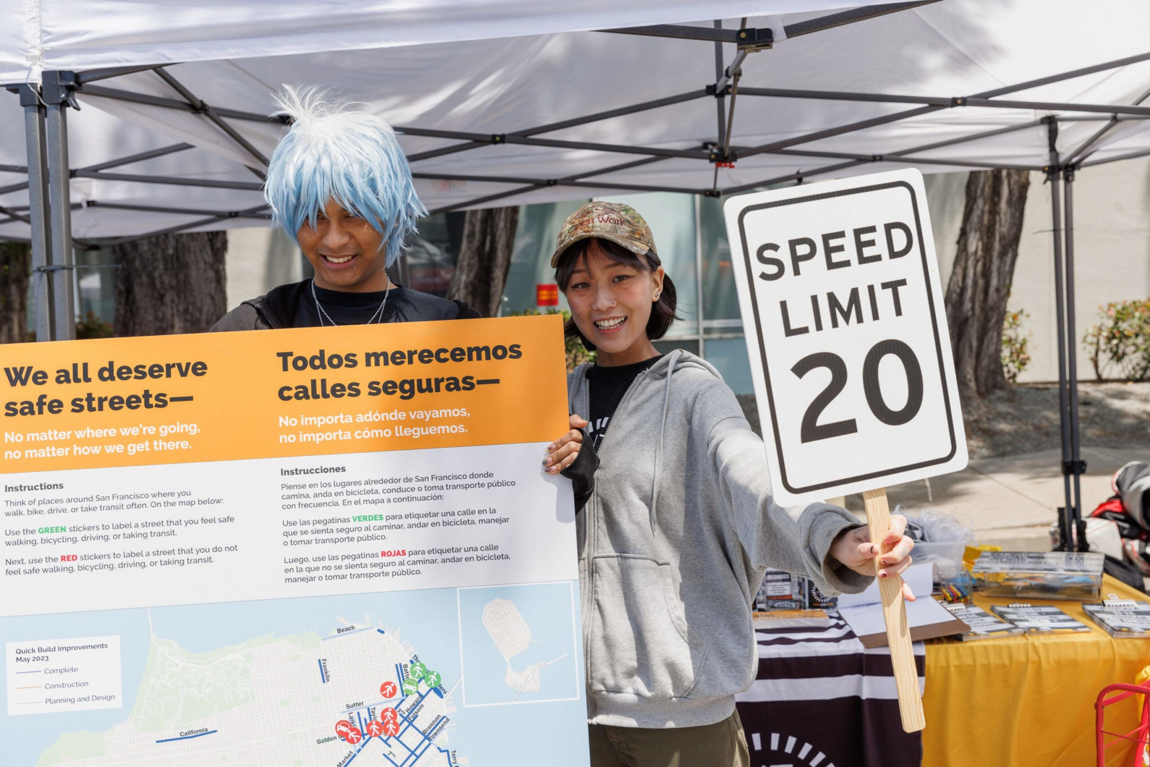 Two people stand by a booth advocating for safe streets, holding a signboard and a &quot;Speed Limit 20&quot; placard.