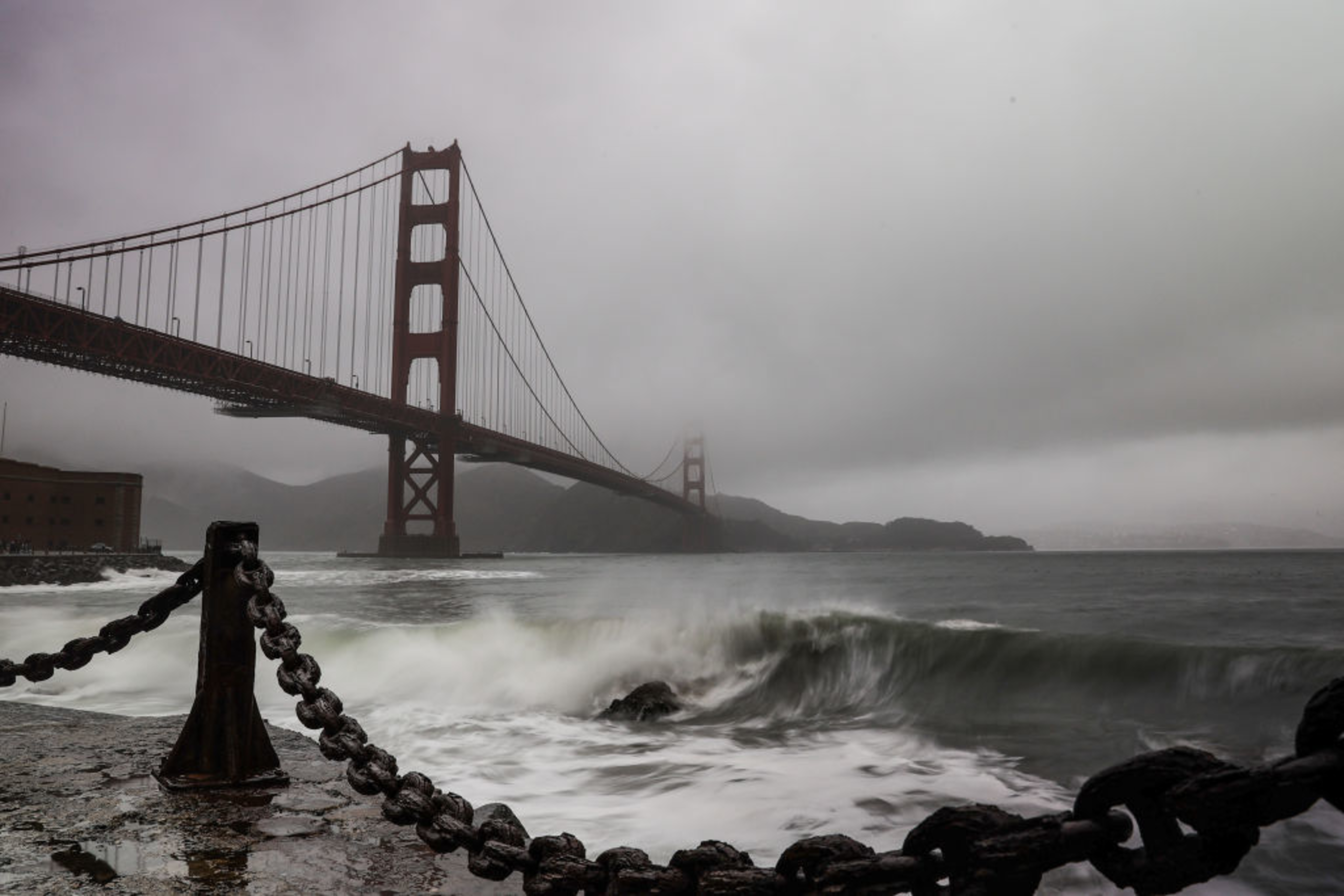 A foggy scene of the Golden Gate Bridge with waves crashing against a rocky shore, linked by a thick chain railing in the foreground.