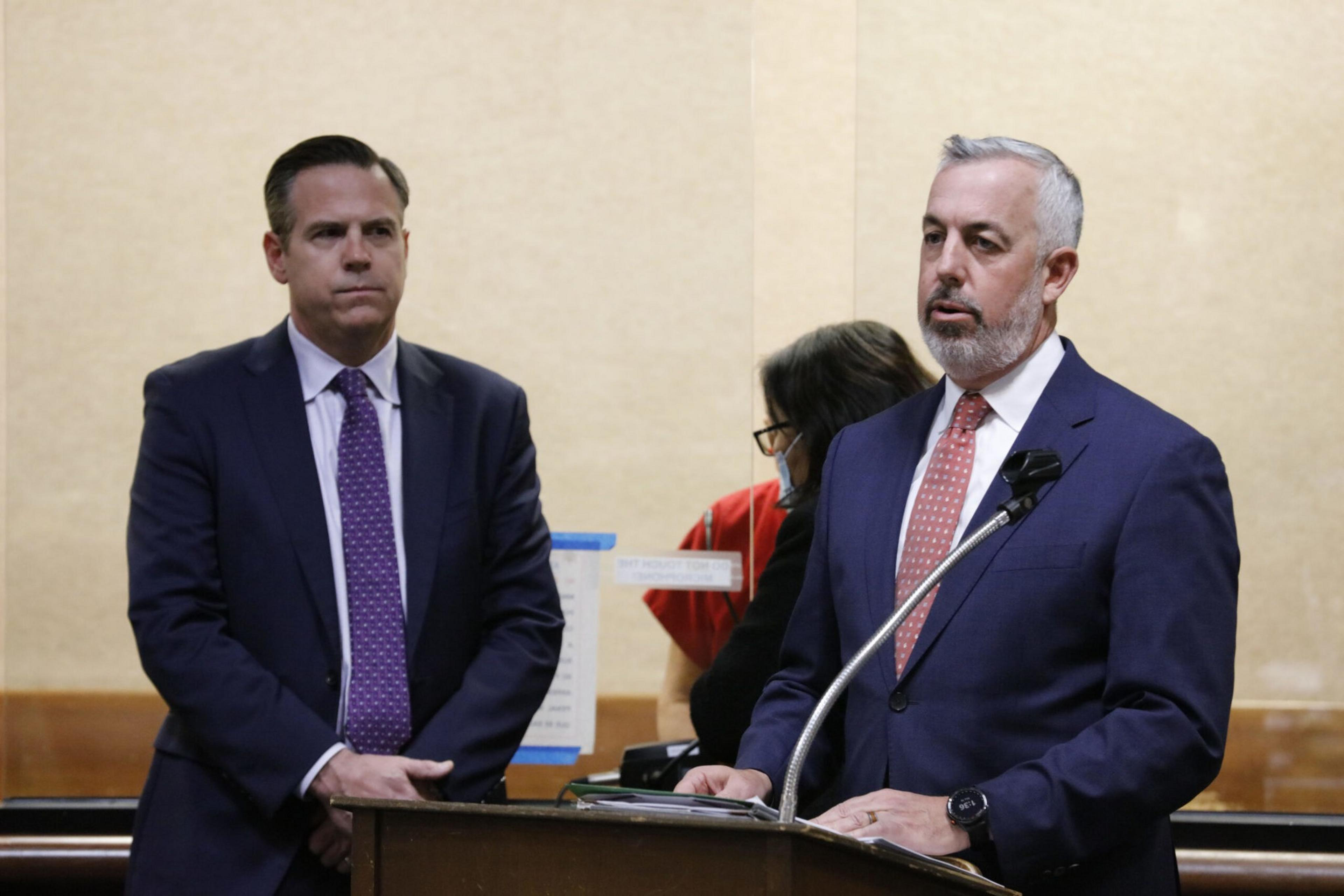 Two men in suits stand at a podium; one speaks into a microphone while the other listens, with a woman in red and black behind a clear barrier.