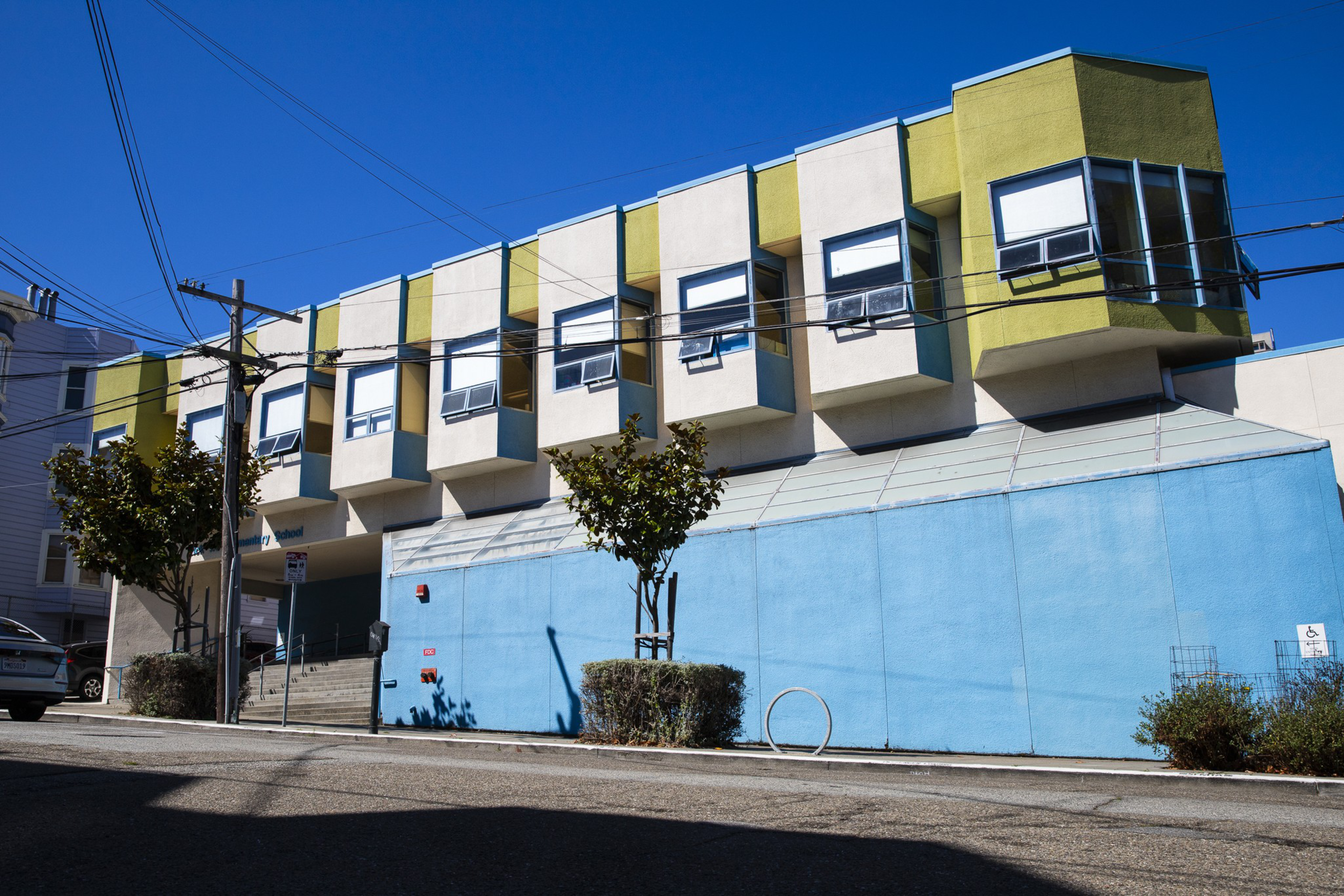 A building with large blue lower walls and upper sections featuring protruding windows painted white and yellow sits under a clear blue sky beside a street.