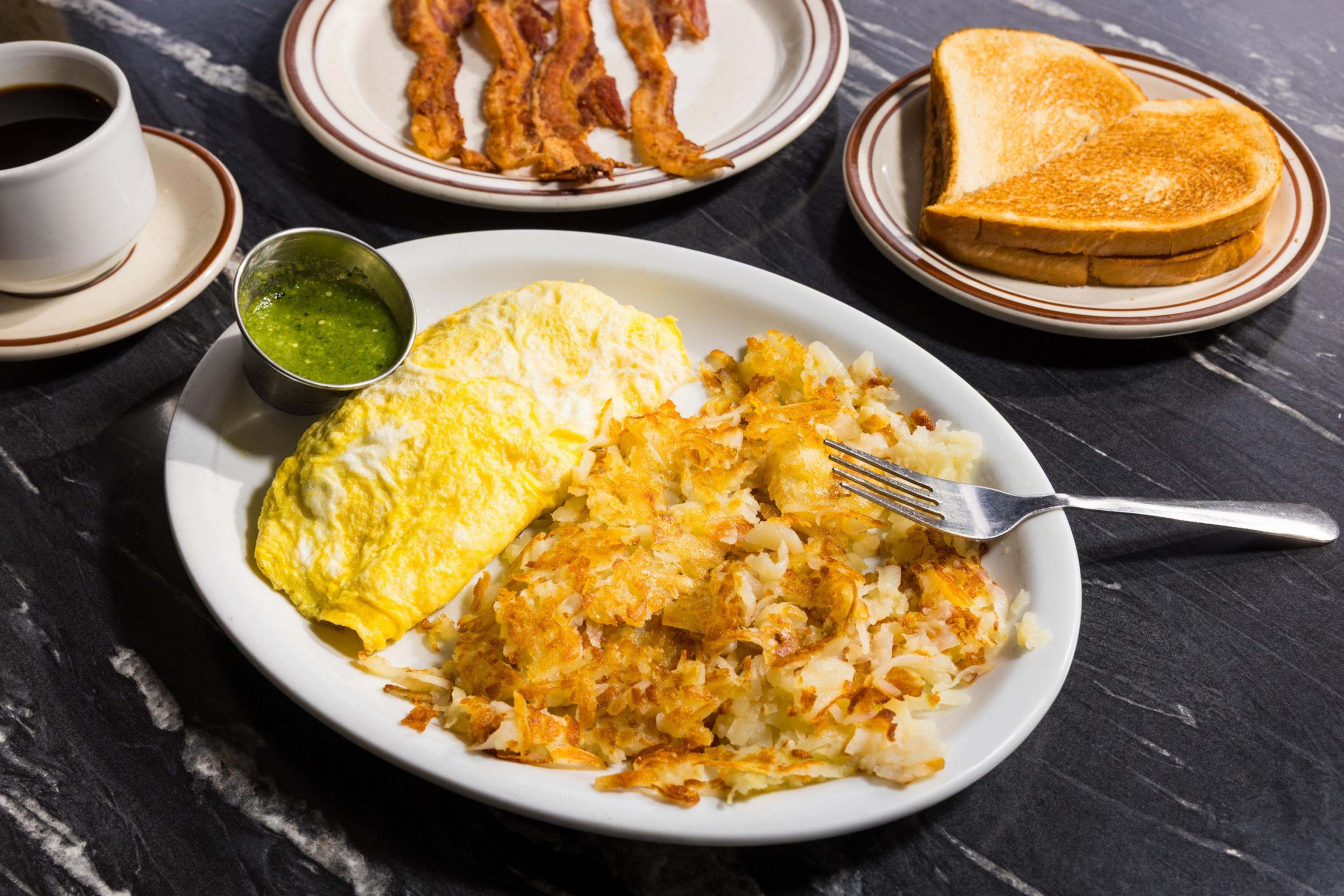 A breakfast spread includes an omelet with golden hash browns, a side of green sauce, bacon strips, toast, and a cup of black coffee on a dark surface.