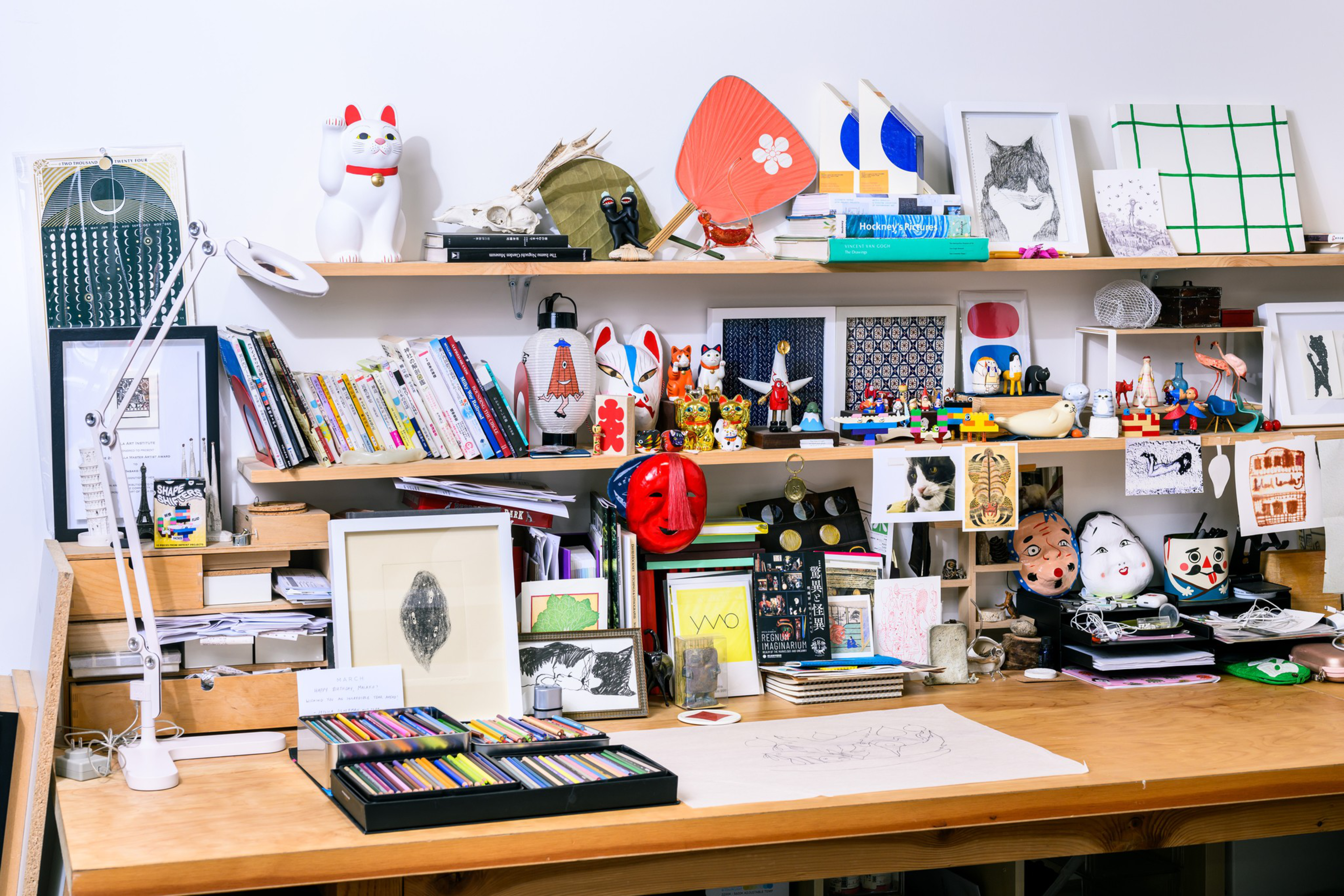 A cluttered desk features books, colorful art supplies, framed pictures, and various quirky collectibles, including figurines and masks, on shelves above.