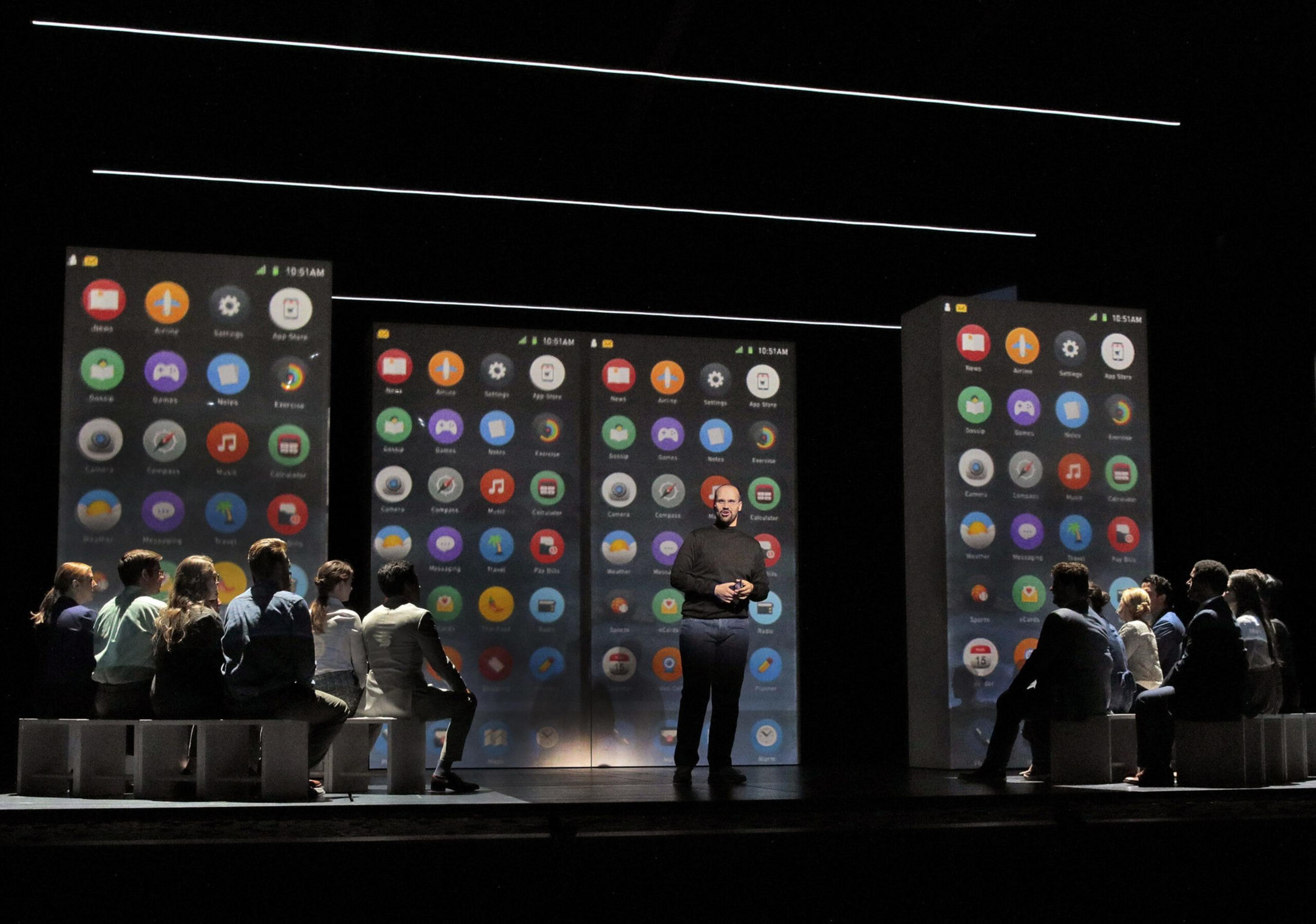 A presenter stands between two groups seated on benches, with three large screens behind showing colorful app icons on a smartphone interface.