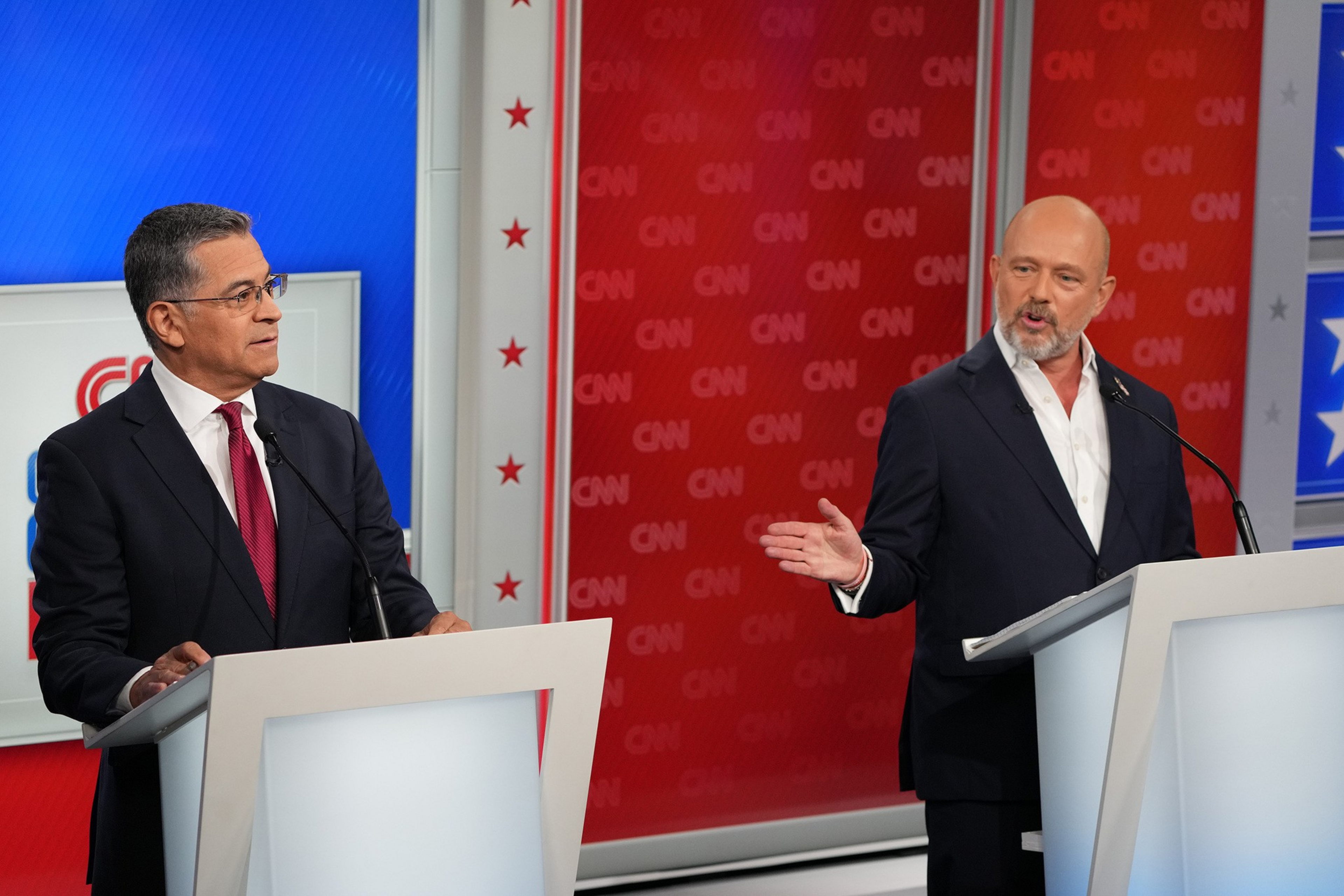 Two men in suits stand behind white podiums with microphones, engaging in a debate on a CNN stage decorated with red, white, and blue colors.