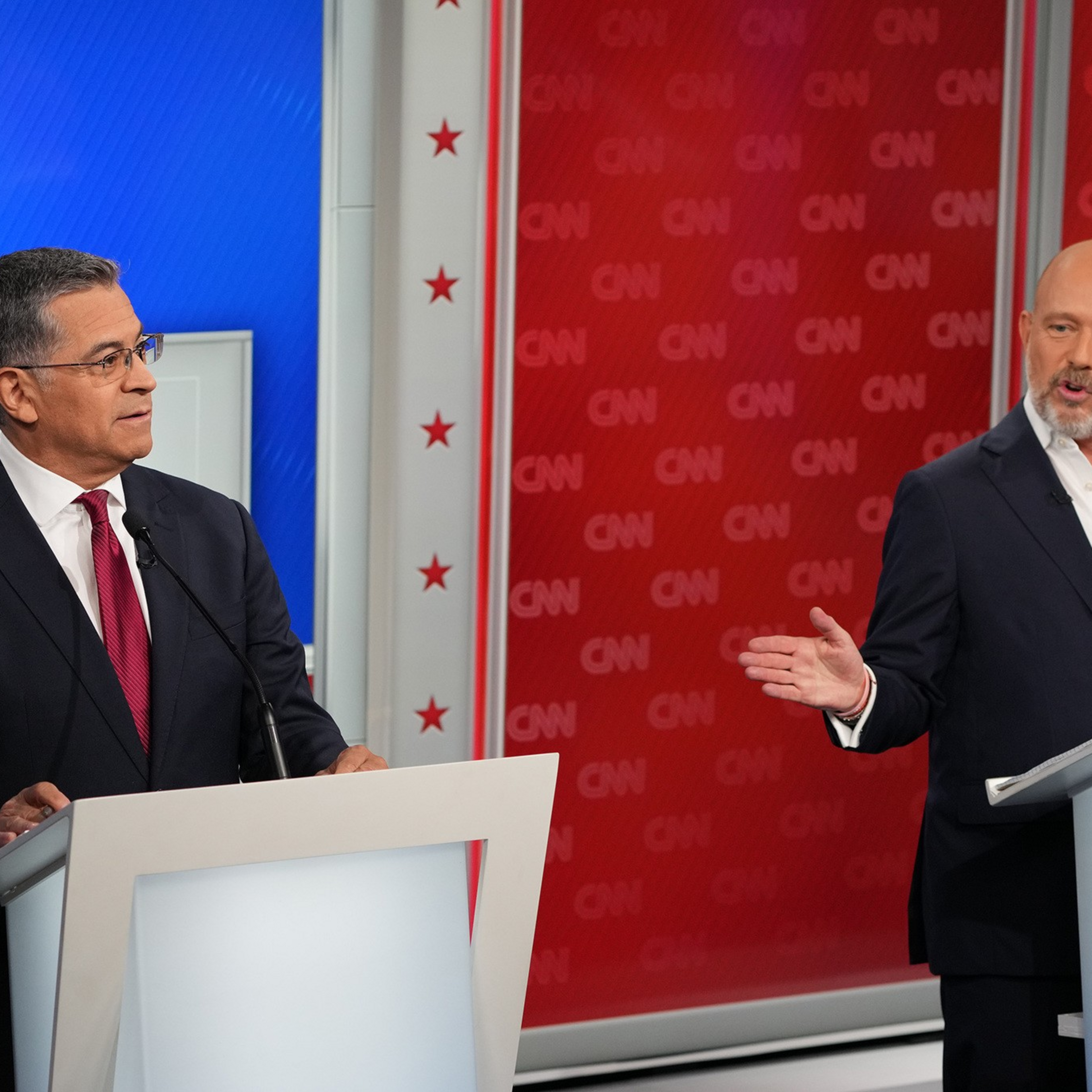 Two men in suits stand behind white podiums with microphones, engaging in a debate on a CNN stage decorated with red, white, and blue colors.