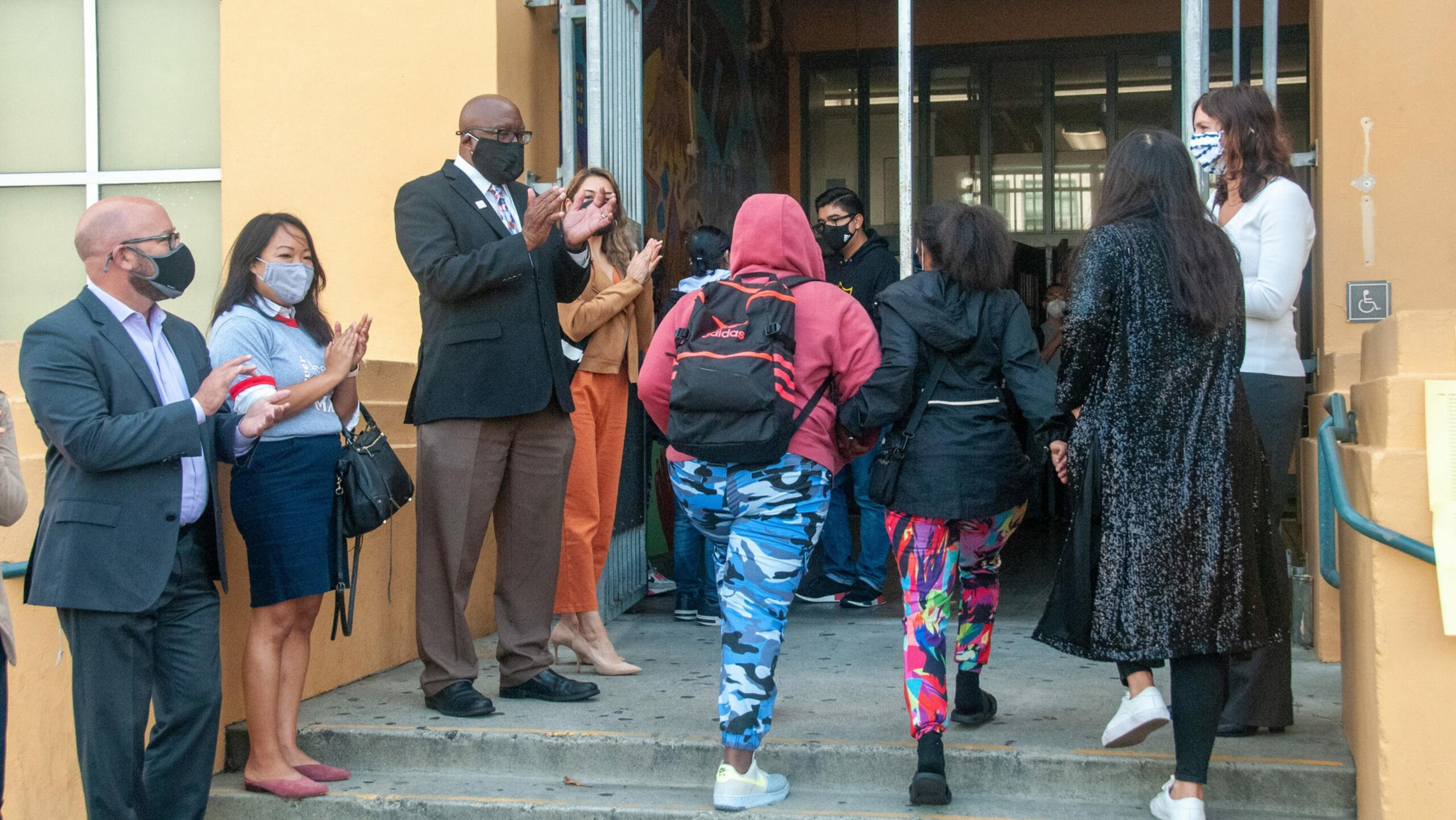 Two students with backpacks walk into a building while adults wearing masks stand outside, clapping and greeting them.