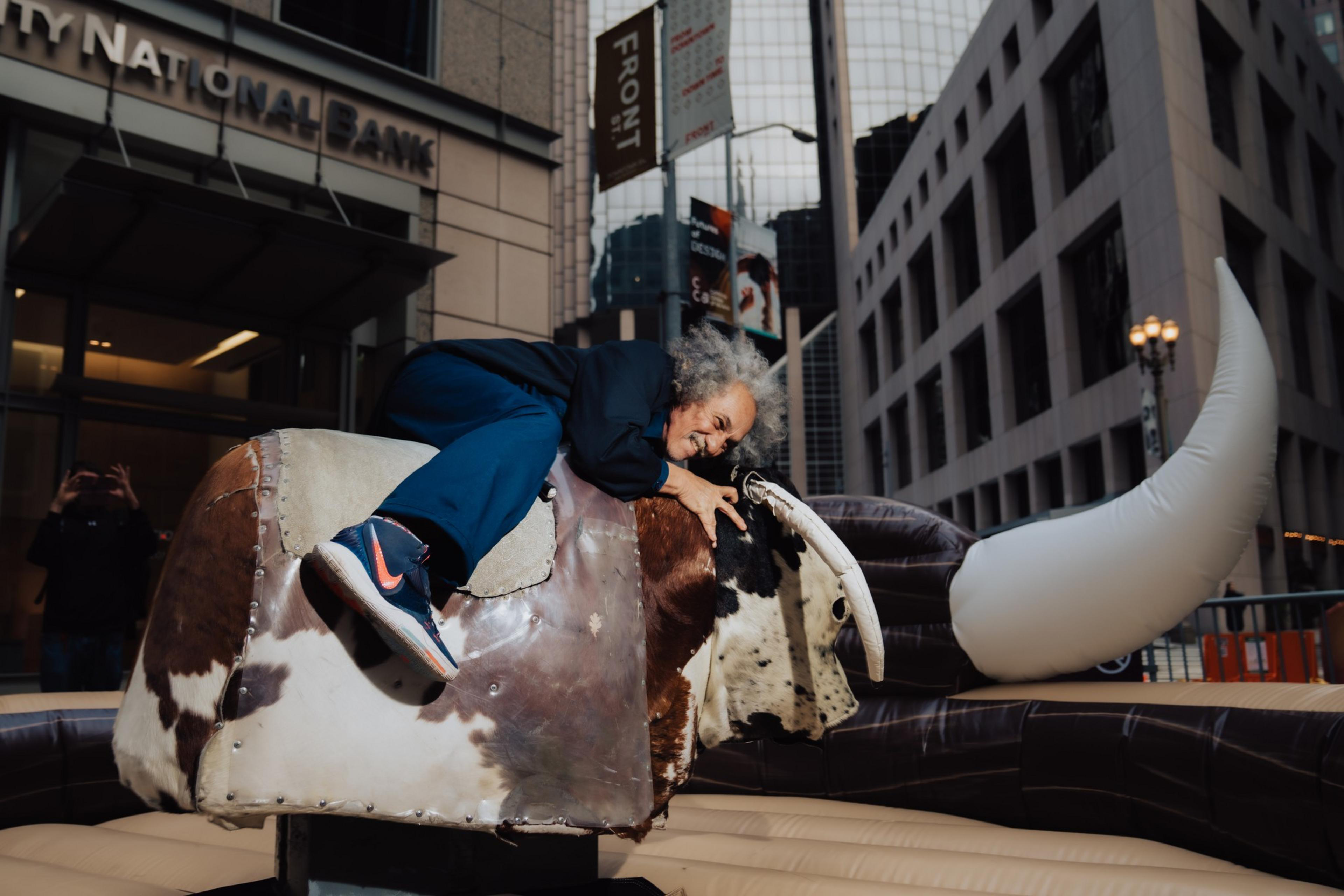 A person with curly hair rides a mechanical bull on a city street, smiling. The background features tall buildings and a sign for City National Bank.