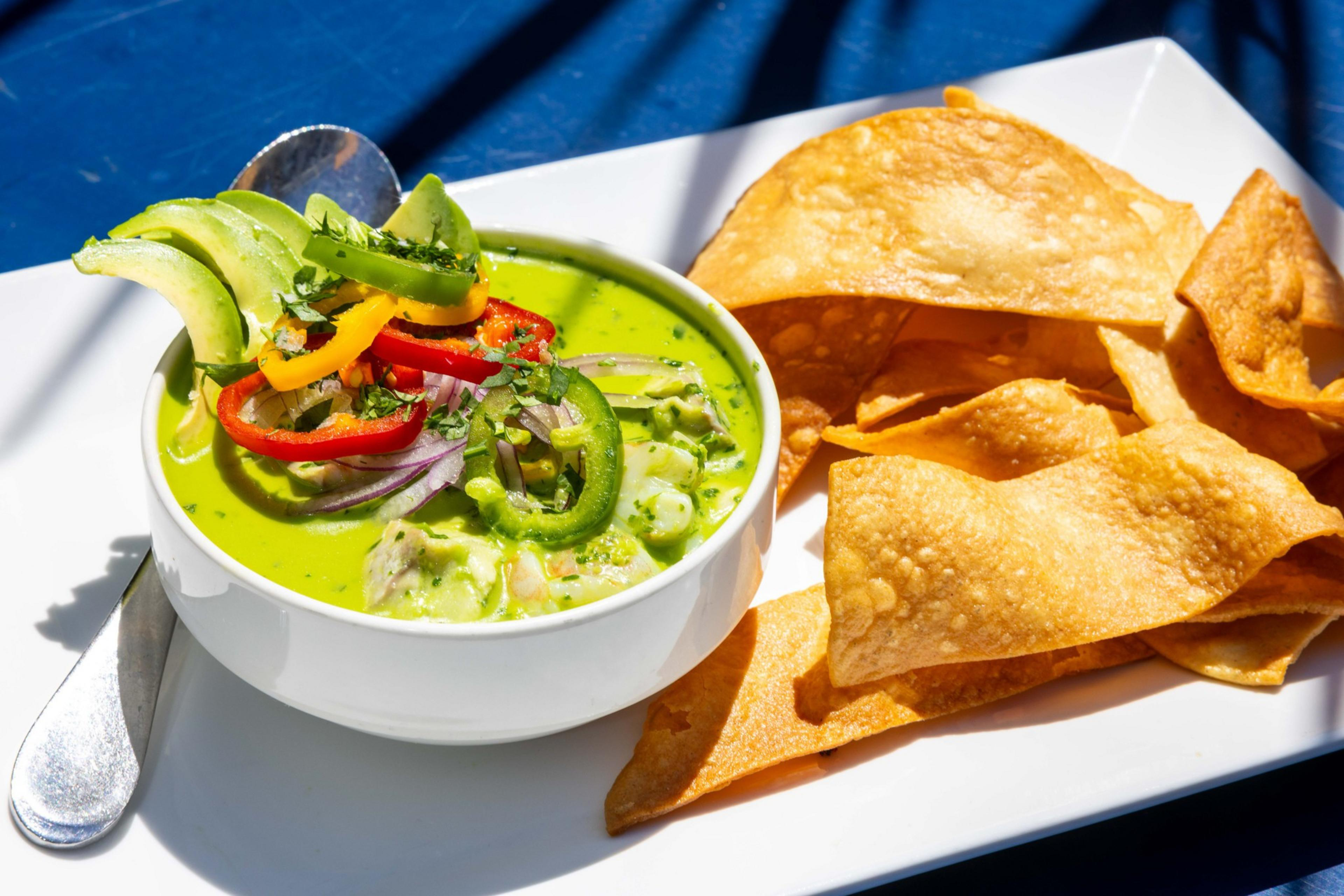 A bowl of green soup topped with avocado slices, red and yellow peppers, and cilantro sits beside crisp tortilla chips on a white rectangular plate.