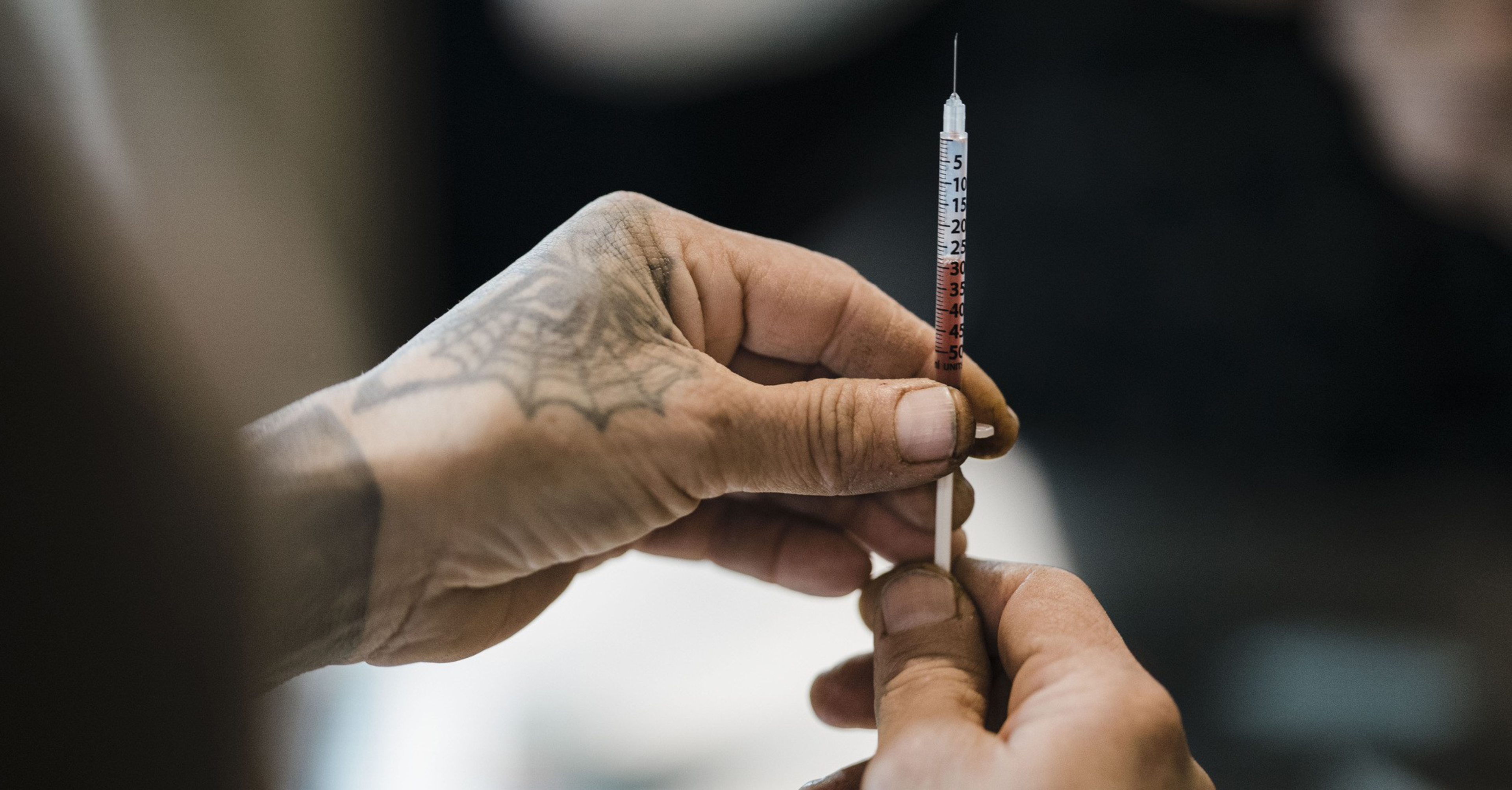 A person with a tattooed hand holds a syringe filled with a red liquid. The background is blurred, focusing attention on the syringe and hands.