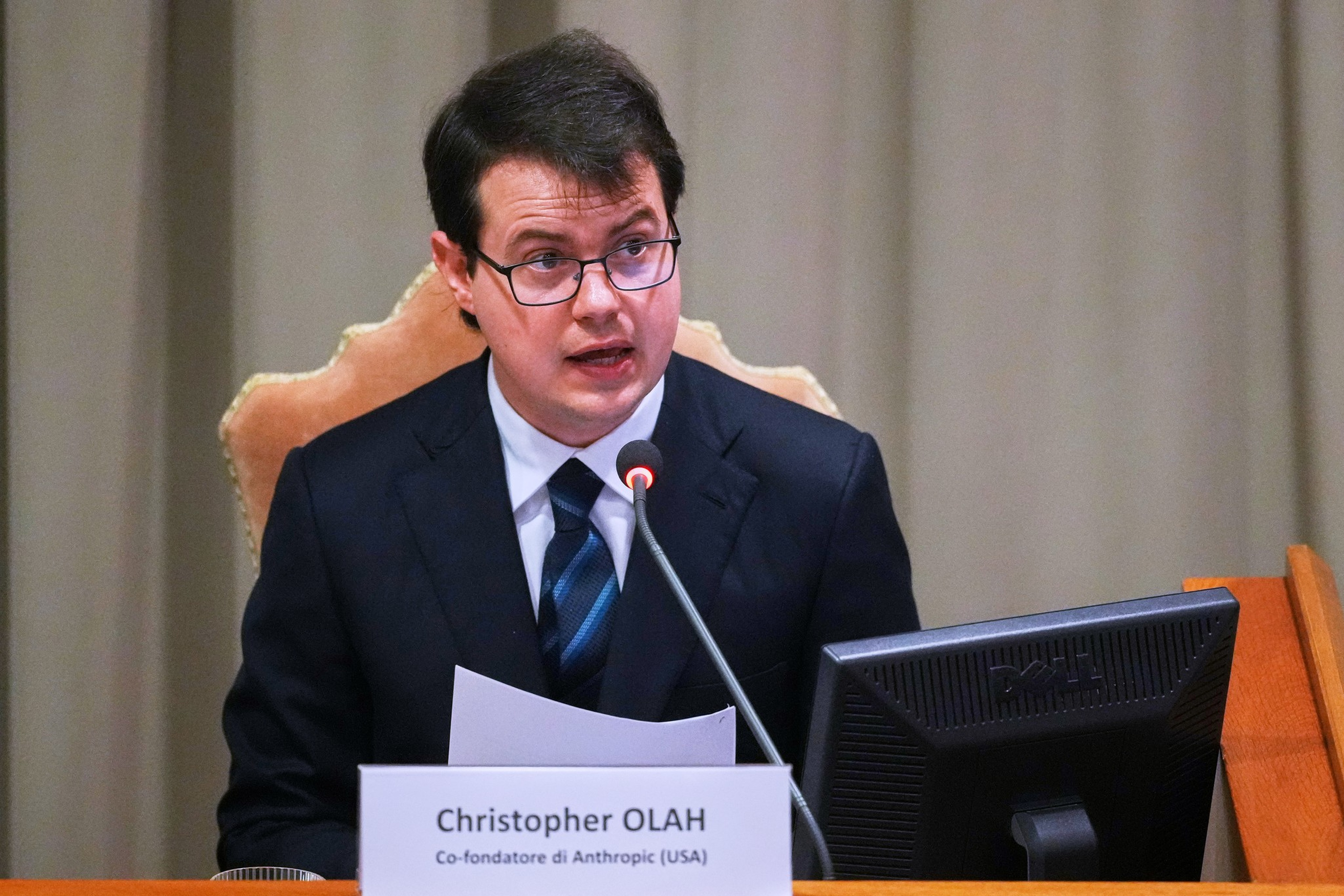 A man in a suit and glasses speaks into a microphone while seated at a desk with a nameplate and computer monitor.