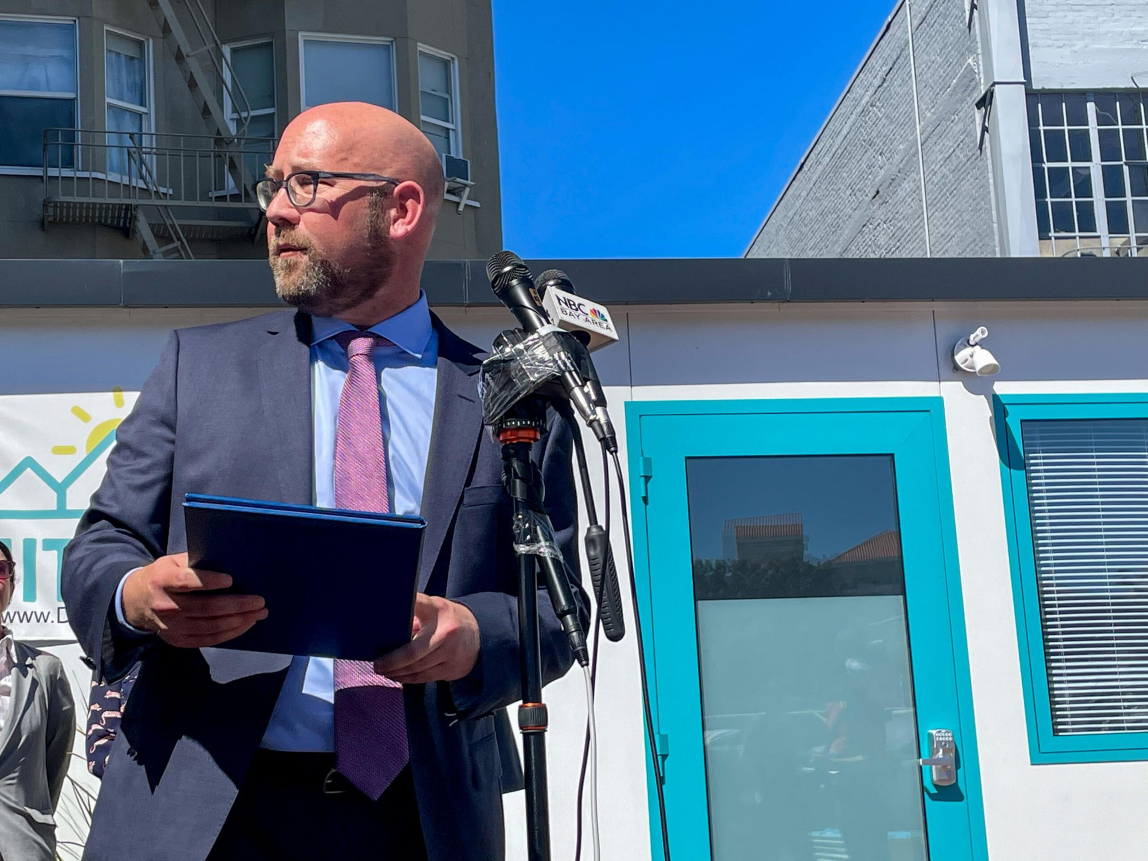 A man in a suit and pink tie stands holding a folder near a microphone with an NBC logo, in front of a building with bright blue doors.