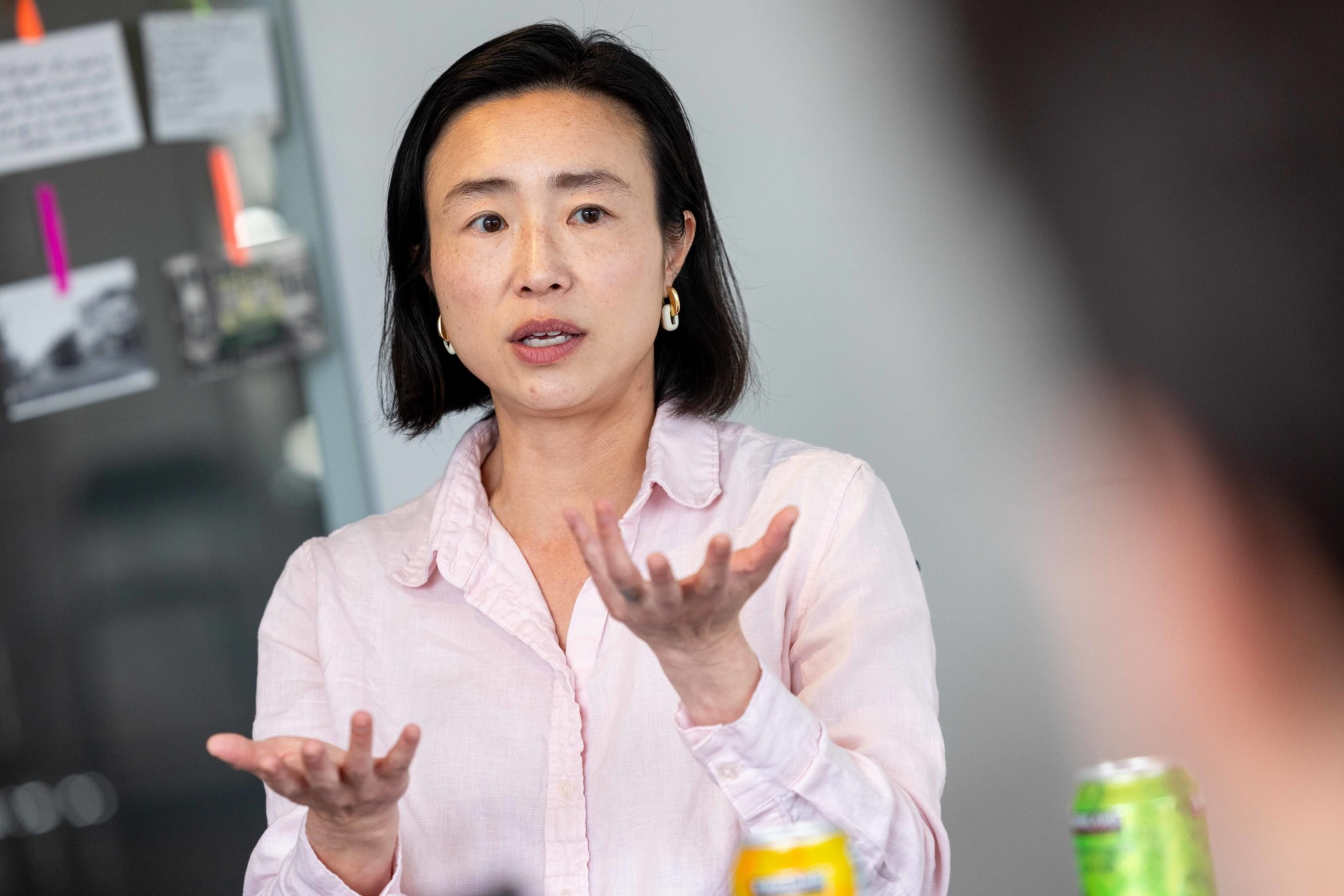 A woman with straight dark hair and gold hoop earrings wears a light pink shirt and gestures with her hands while speaking indoors.
