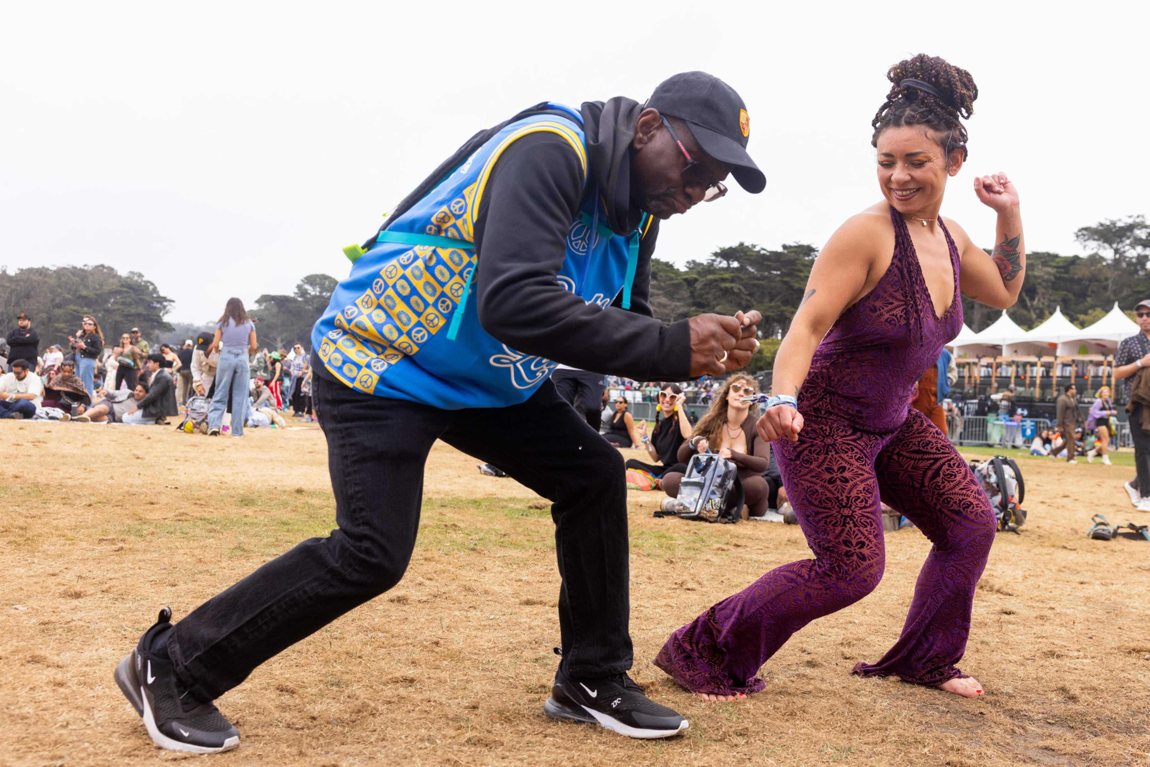 A man in a blue hoodie and cap and a woman in a purple jumpsuit dance energetically on grass with a crowd watching behind them.