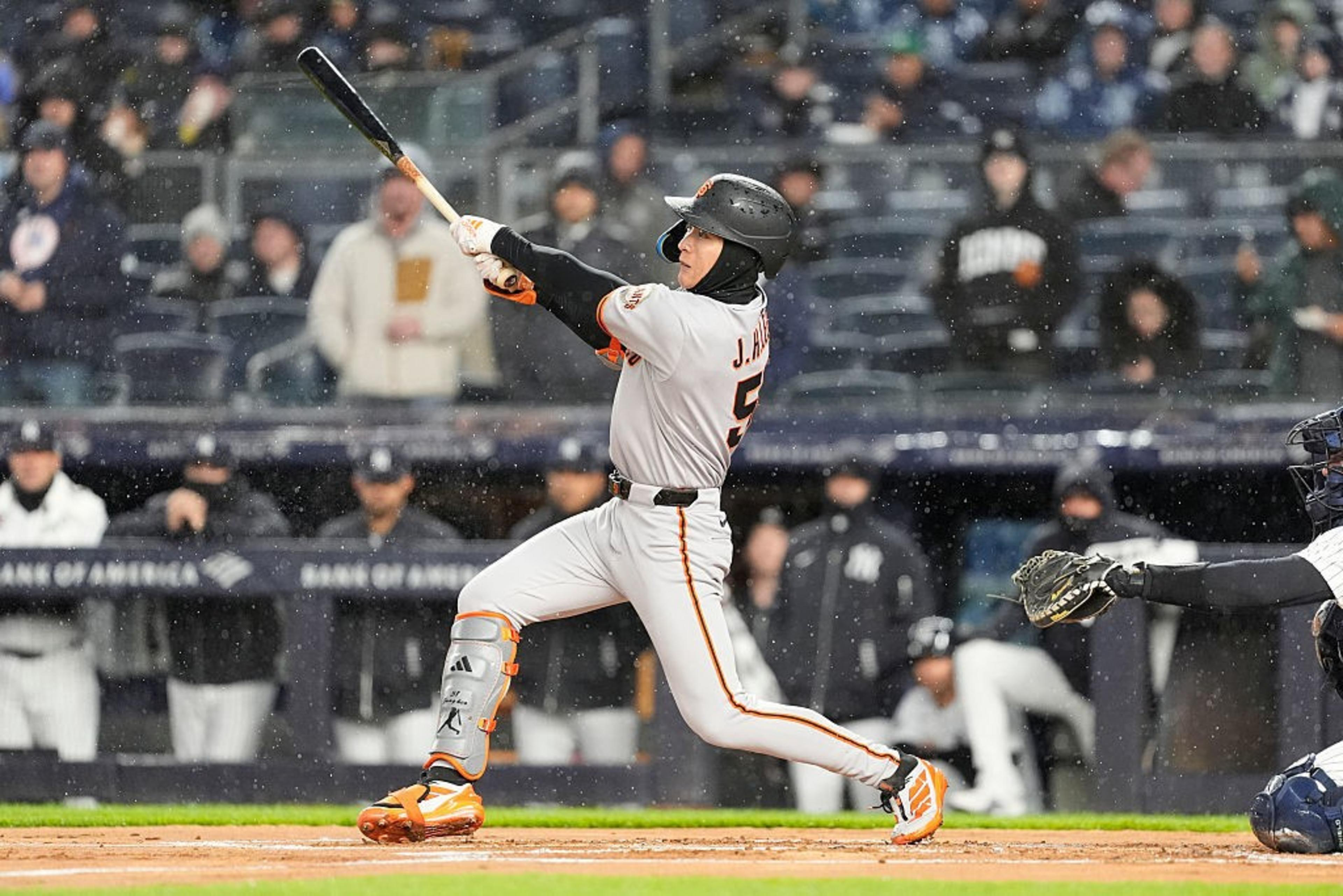 A baseball player in a gray and orange uniform is mid-swing in a rainy stadium. He's wearing a helmet and leg guards, with spectators visible in the background.