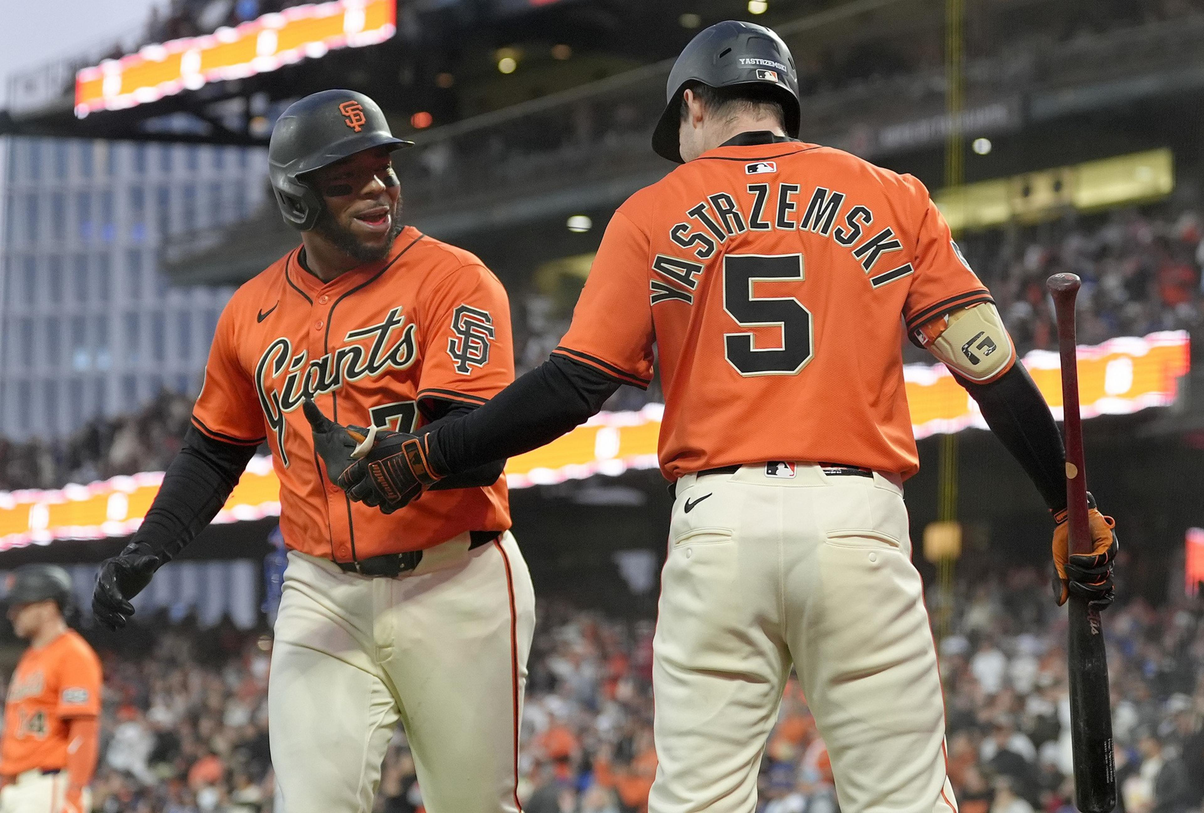 Two baseball players in orange San Francisco Giants jerseys are celebrating on the field. One holds a bat while they appear to exchange a handshake.