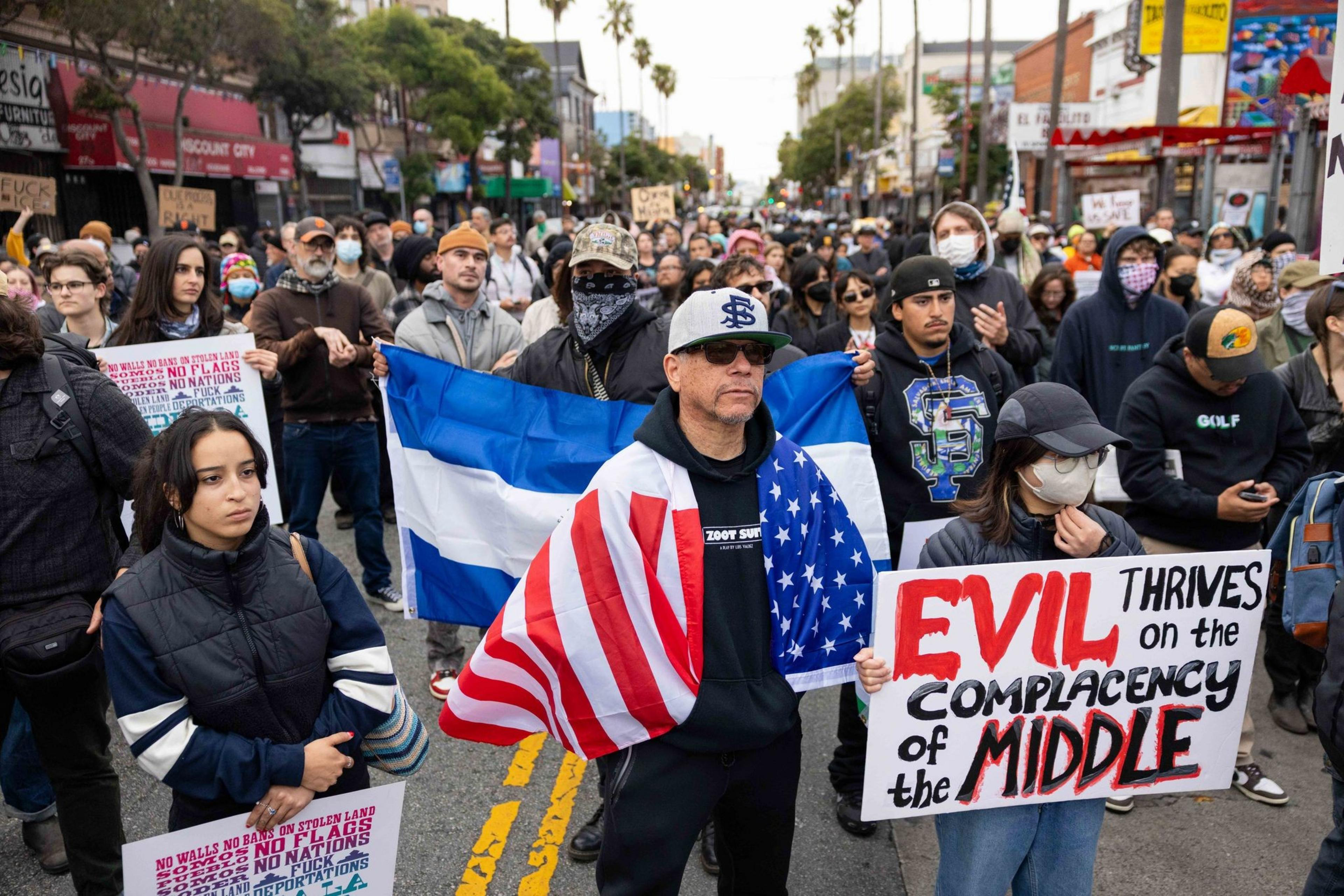 The image shows a diverse group of protesters on a street, holding signs and flags. One person is wrapped in a U.S. flag, and another holds a sign about complacency.