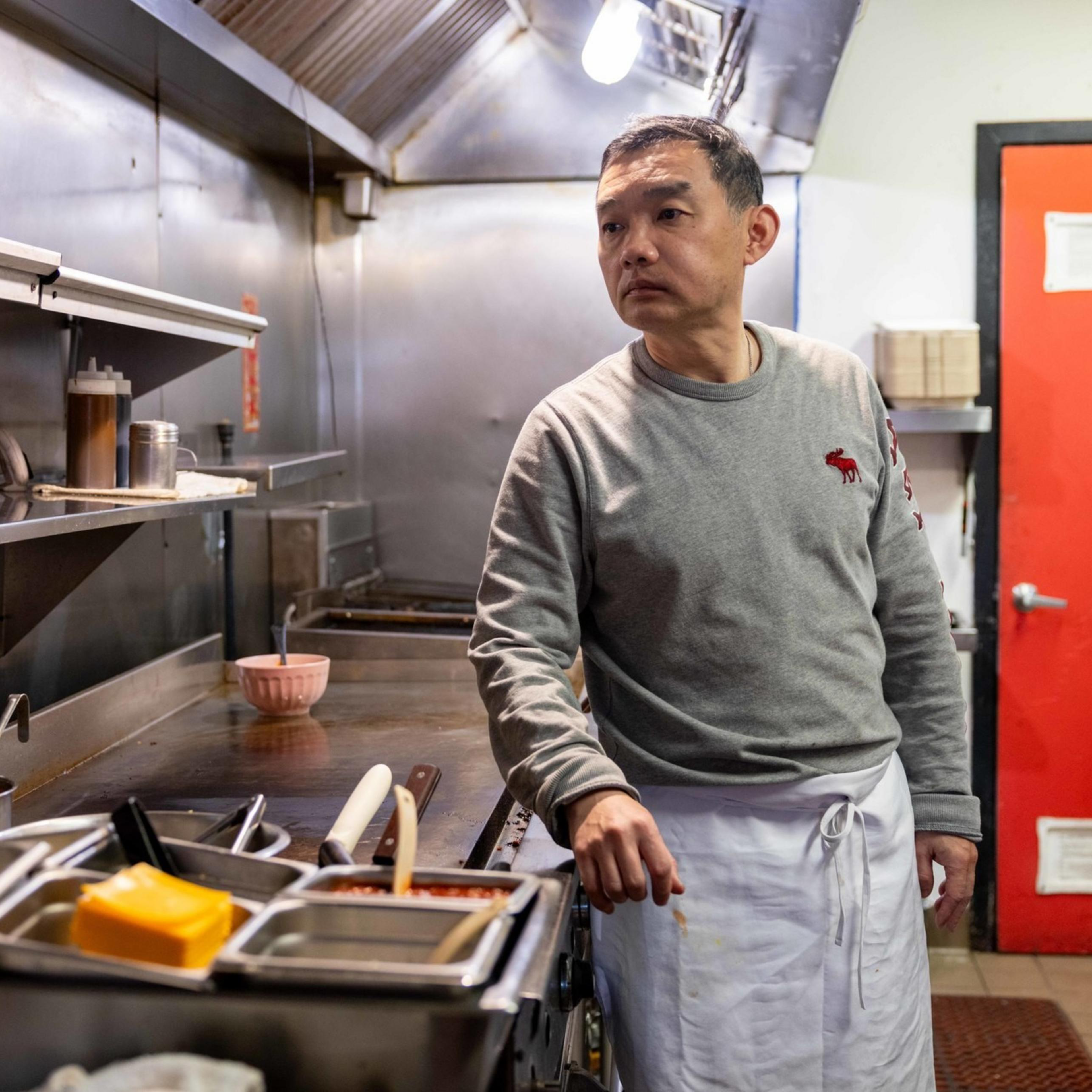 A man in a gray sweater and white apron stands in a commercial kitchen. He looks to the side, with utensils and kitchen equipment around him. A red door is visible.
