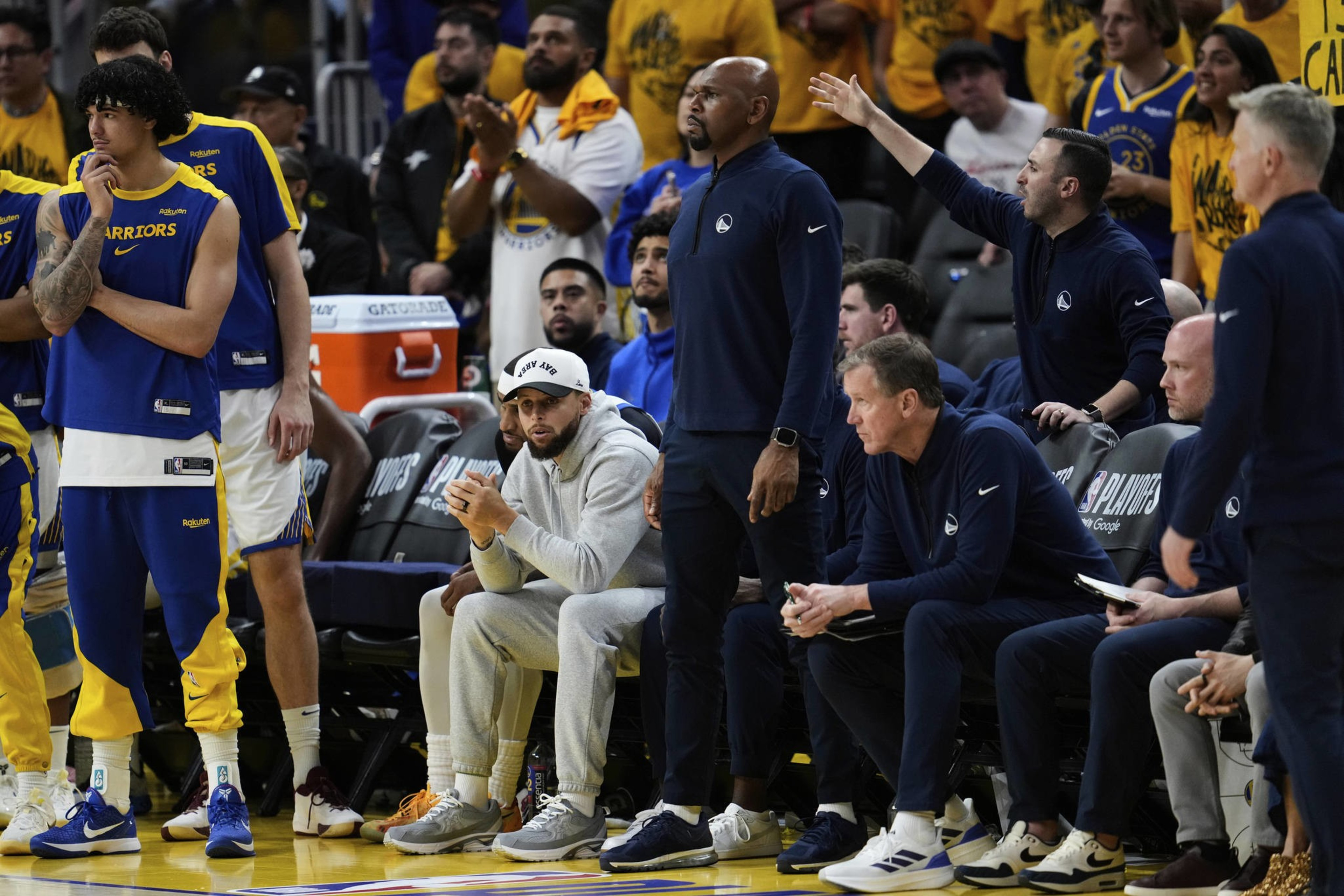 A basketball team watches the game from the sidelines. Players in yellow and blue stand next to coaches in navy. Fans cheer in yellow shirts behind them.