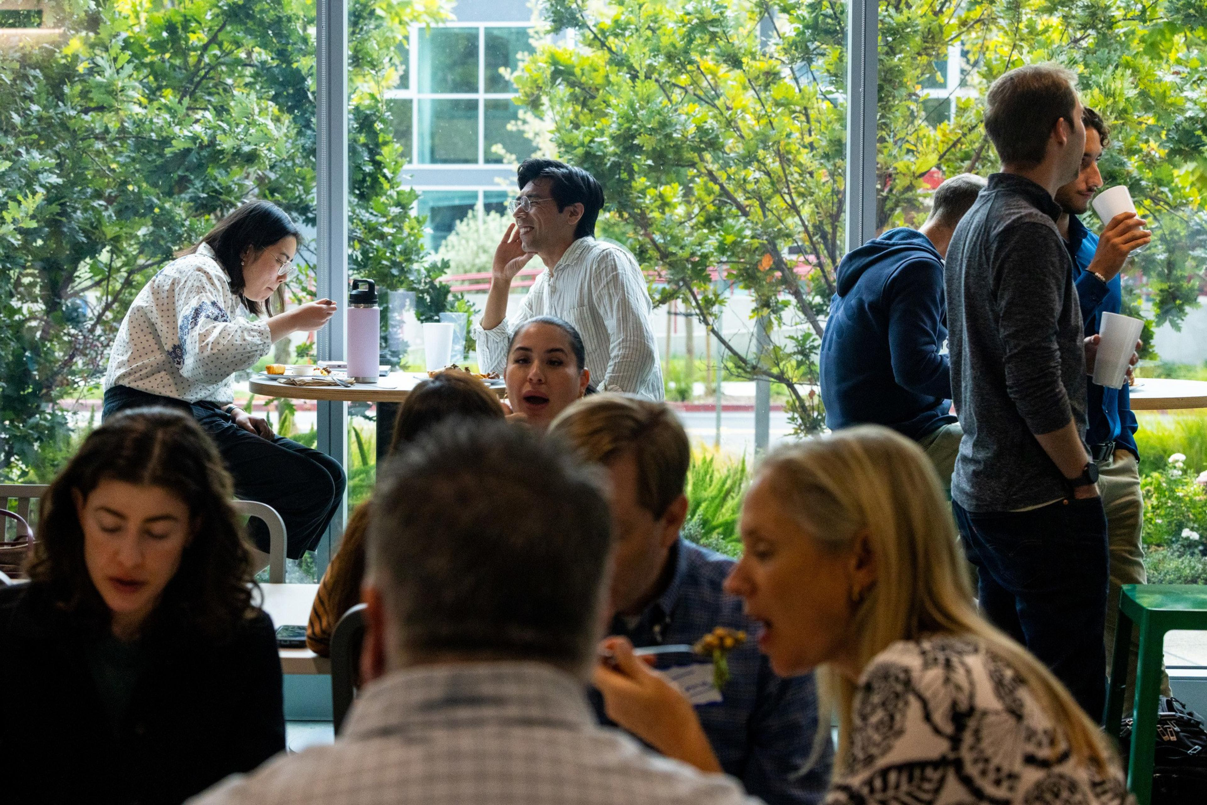 People are eating, drinking, and talking inside a cafe with large windows showing green trees and a building outside.
