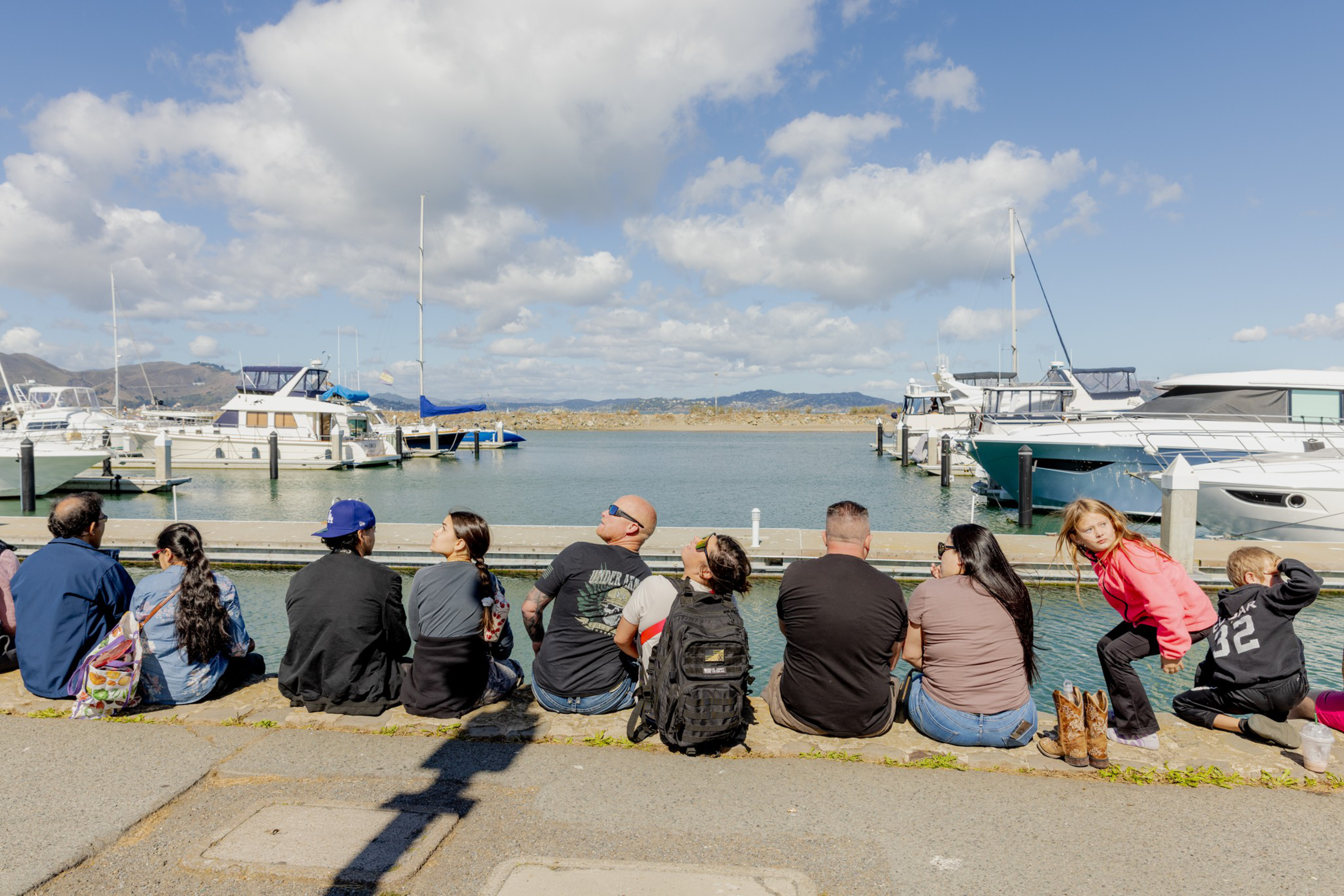 A group of people sit on the ground facing a marina with boats docked, under a partly cloudy sky on a sunny day.