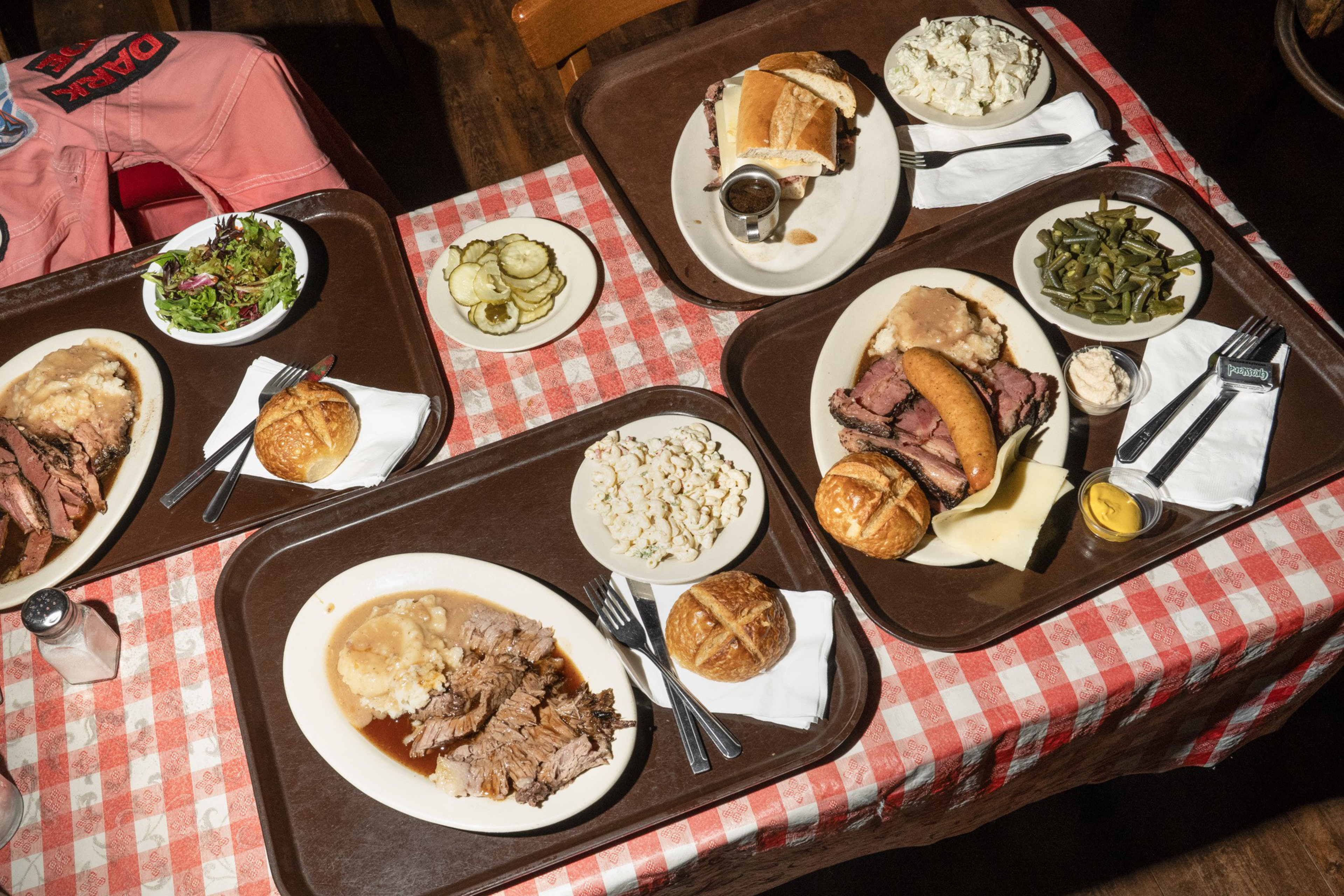Three trays of barbecue meals with brisket, sausage, rolls, mashed potatoes, popcorn, green beans, salad, pickles, and condiments on a red-and-white checkered table.