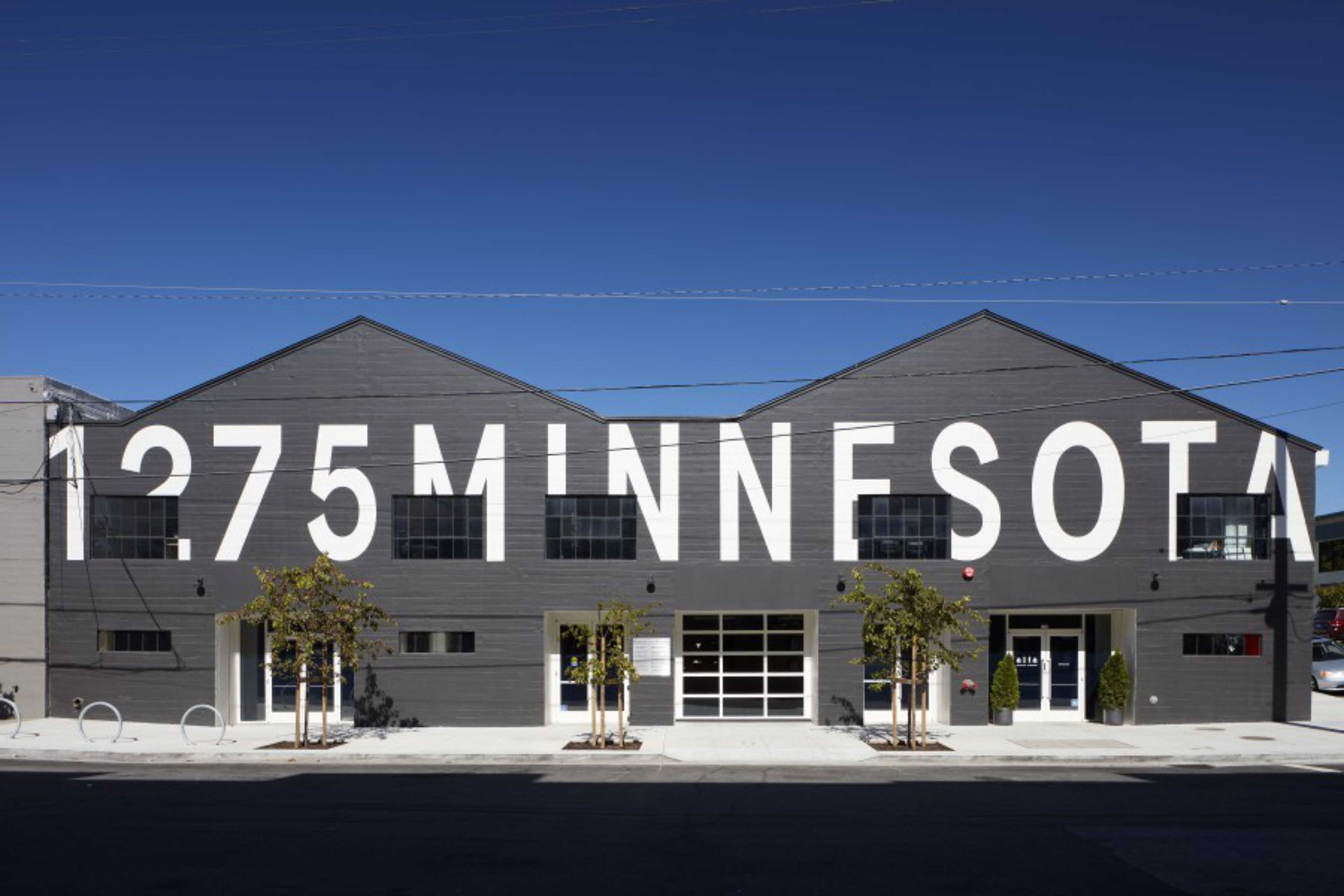 A large dark gray building has big white letters spelling “1275 MINNESOTA” across its facade under a clear blue sky.