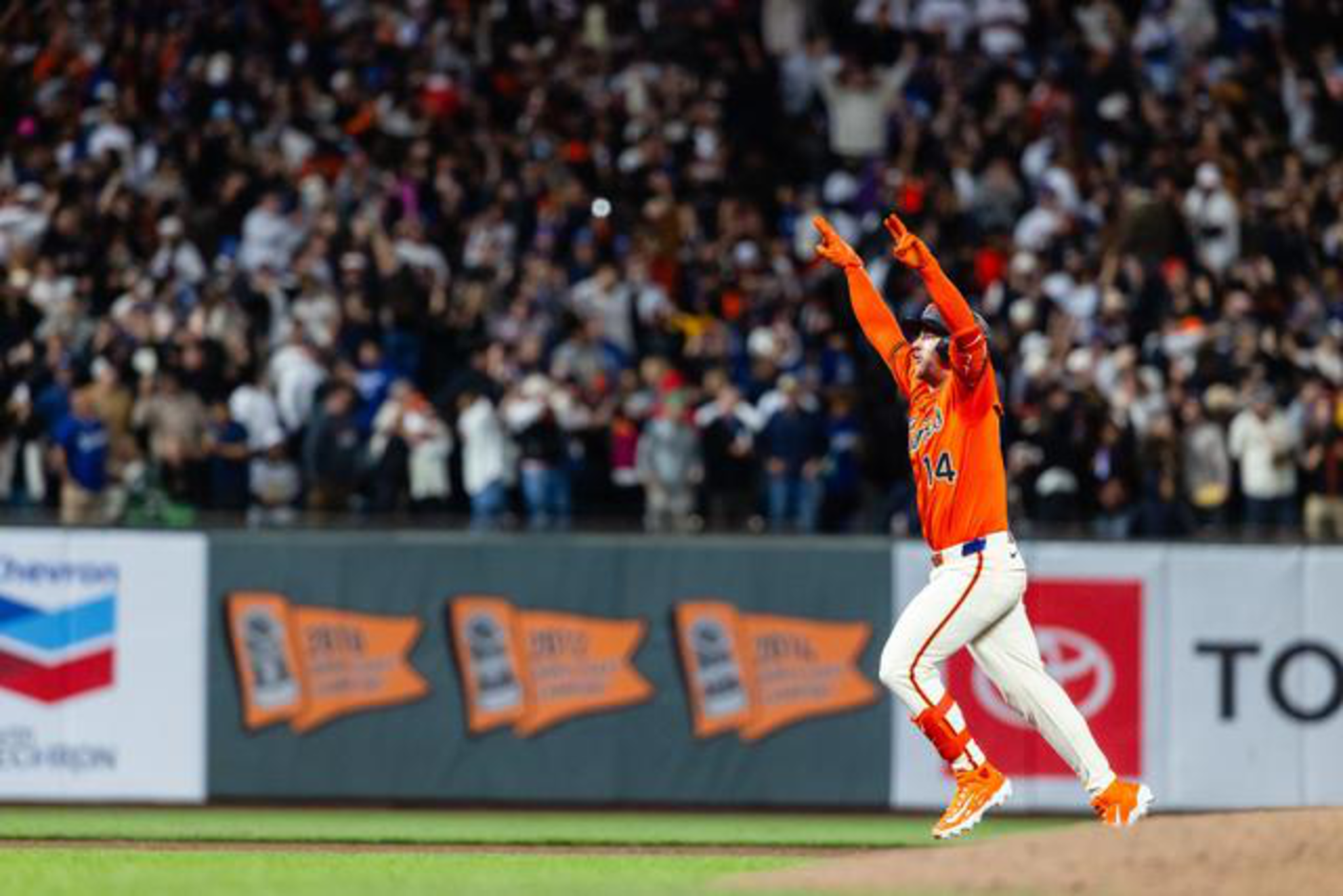 A baseball player in an orange jersey and white pants runs with both arms raised, while a large crowd cheers in the background.