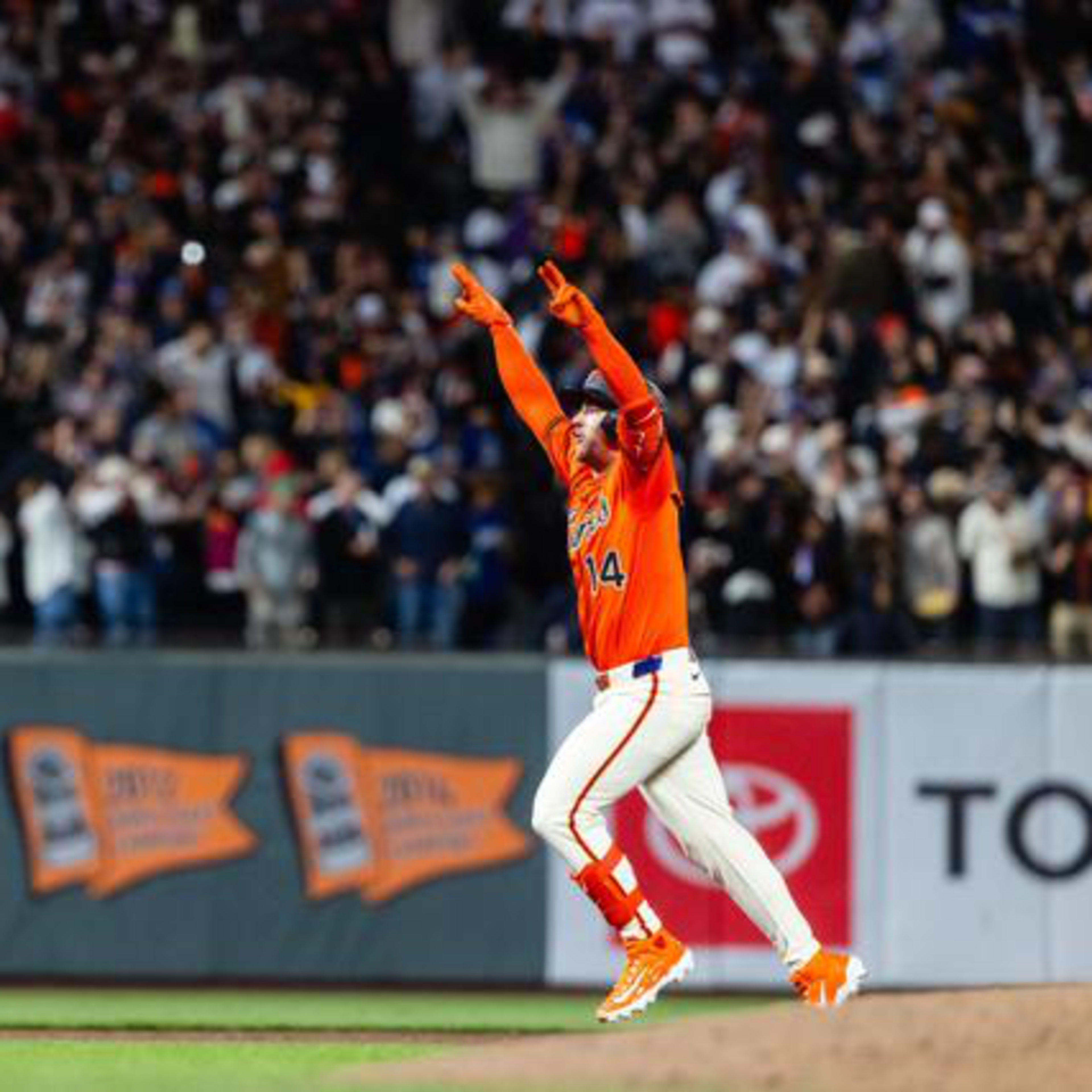 A baseball player in an orange jersey and white pants runs with both arms raised, while a large crowd cheers in the background.