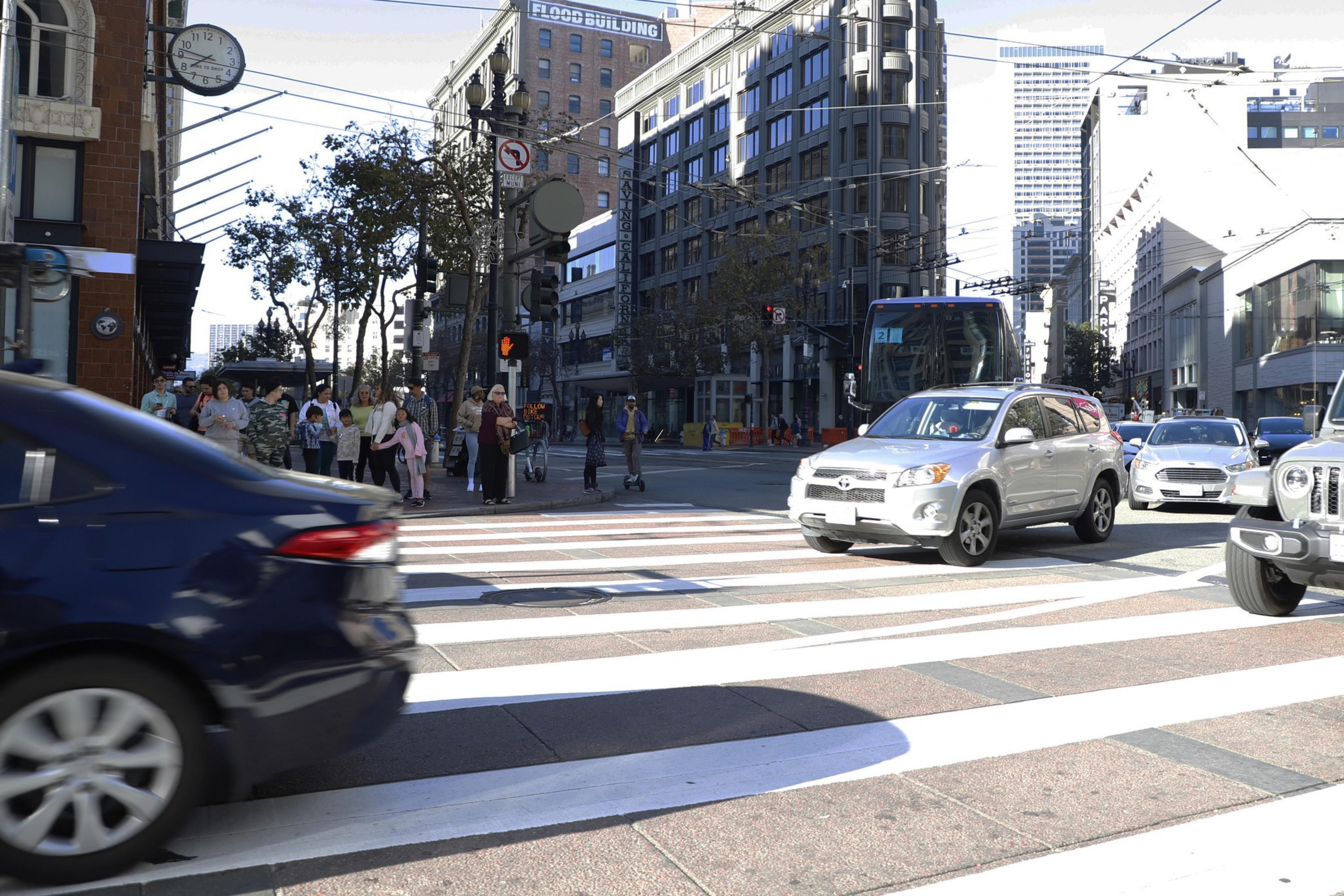 Several cars drive through an intersection as people wait on the corner to cross the street.