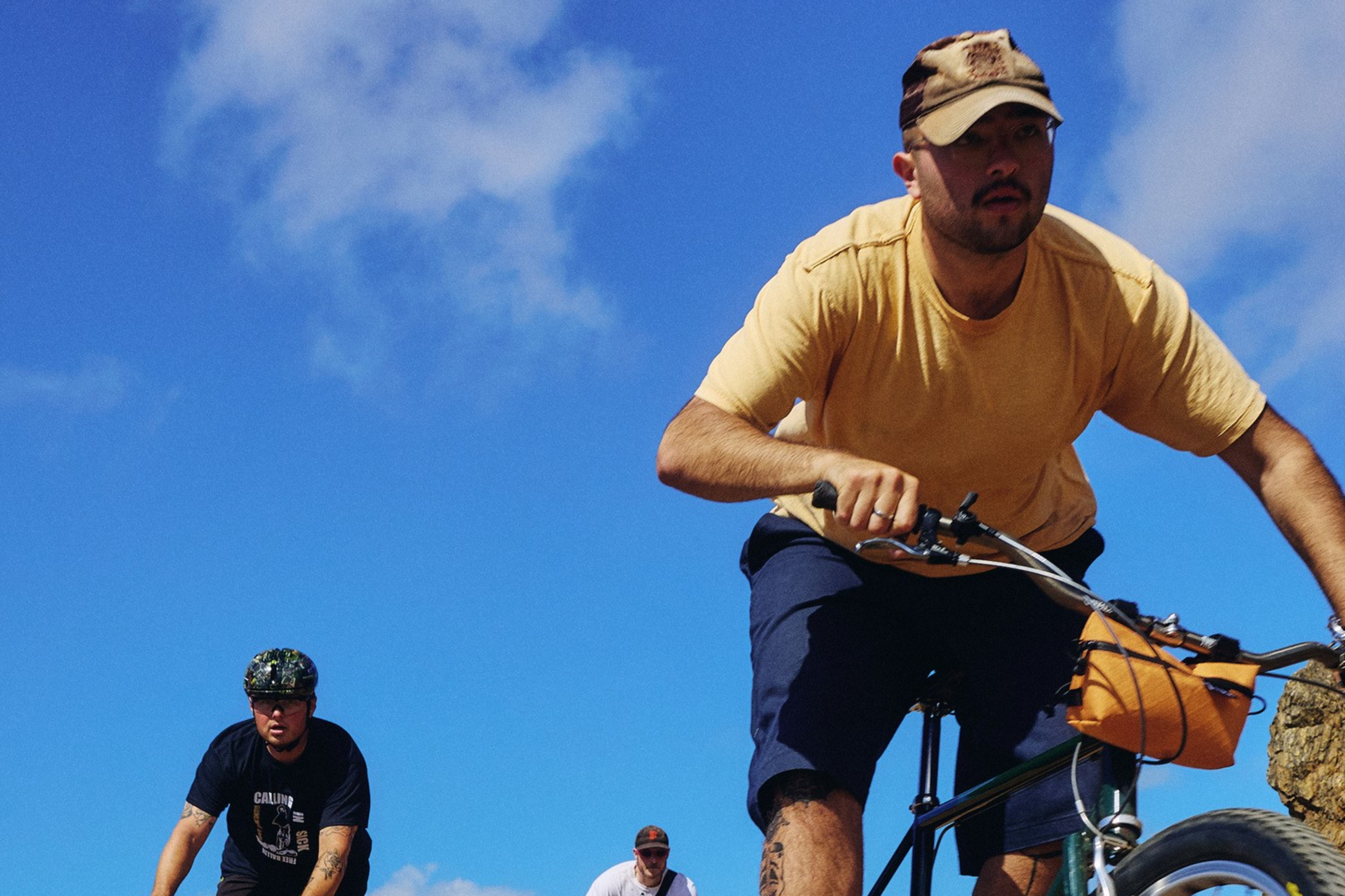 Three men ride bicycles on a sunny dirt trail with a clear blue sky and hills in the background.
